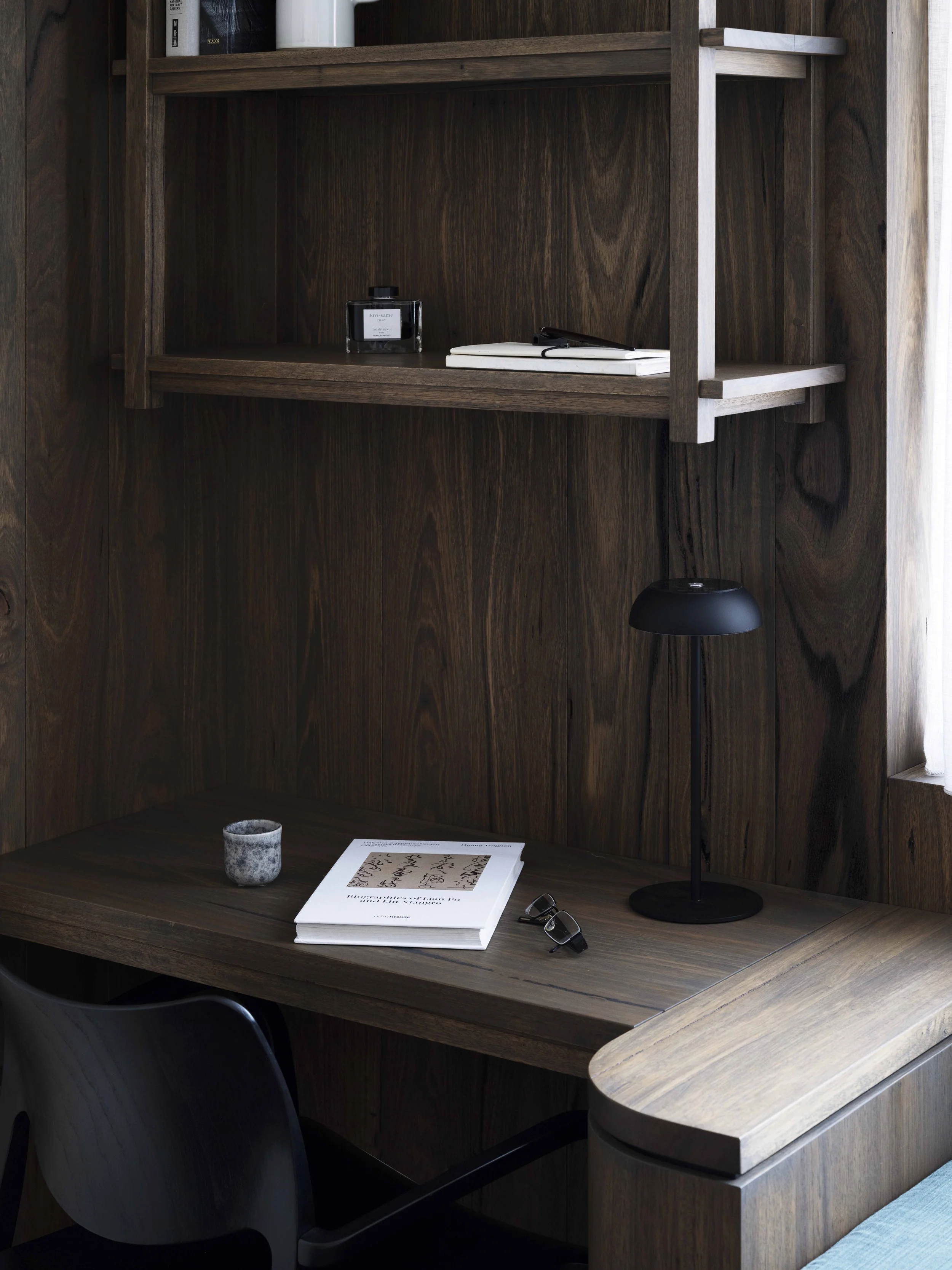 A wooden desk with a black desk lamp, a pair of glasses, a book, a small cup, and a magazine. There are shelves above the desk with a black ink bottle, a small stack of books, and a white cylindrical object. The wall and furniture are made of dark wood, and a window with a curtain is partially visible on the right.