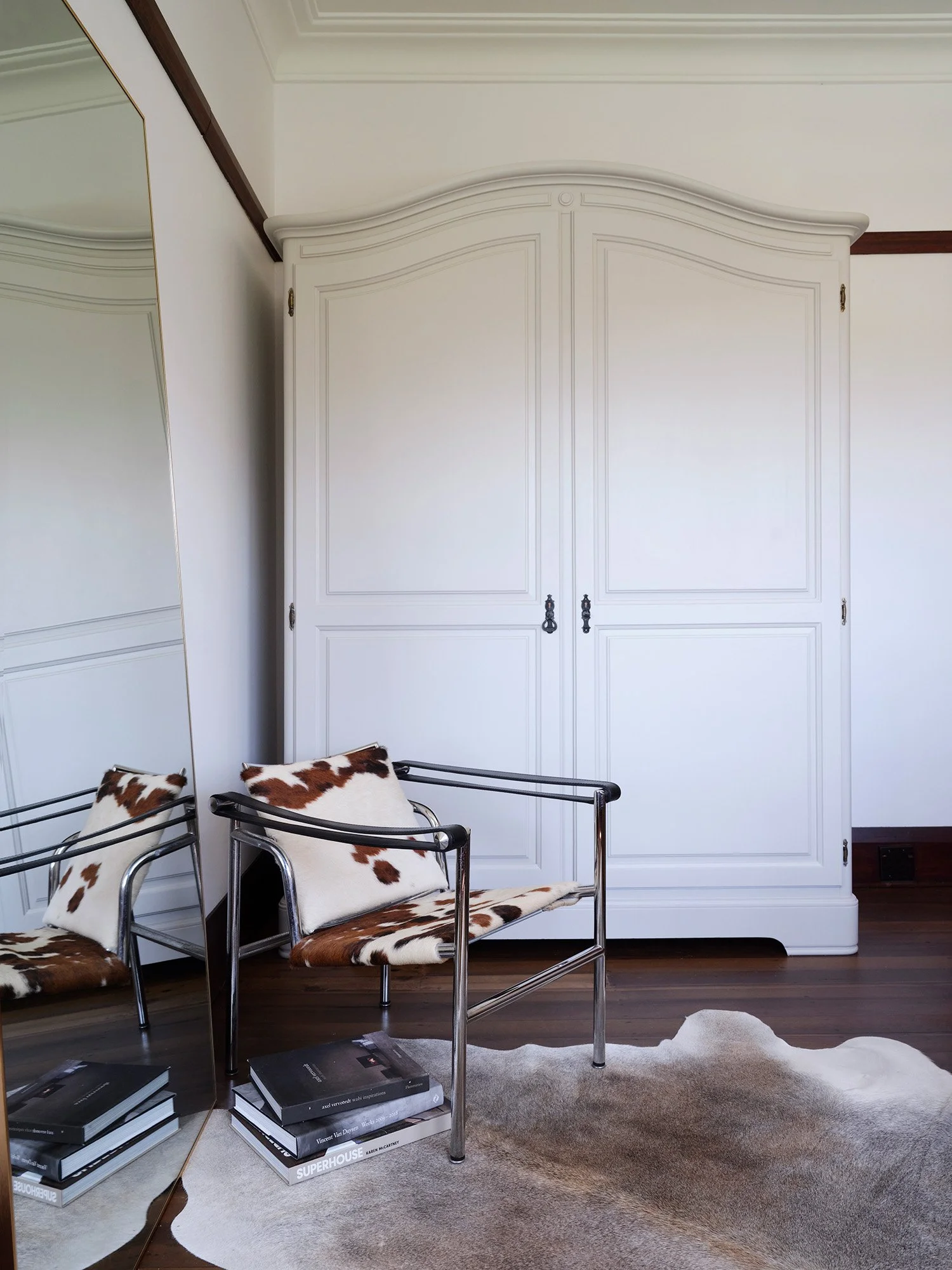 A room with a white wardrobe, a full-length mirror, two cowhide pattern chairs with pillows, a cowhide rug, and stacked books.