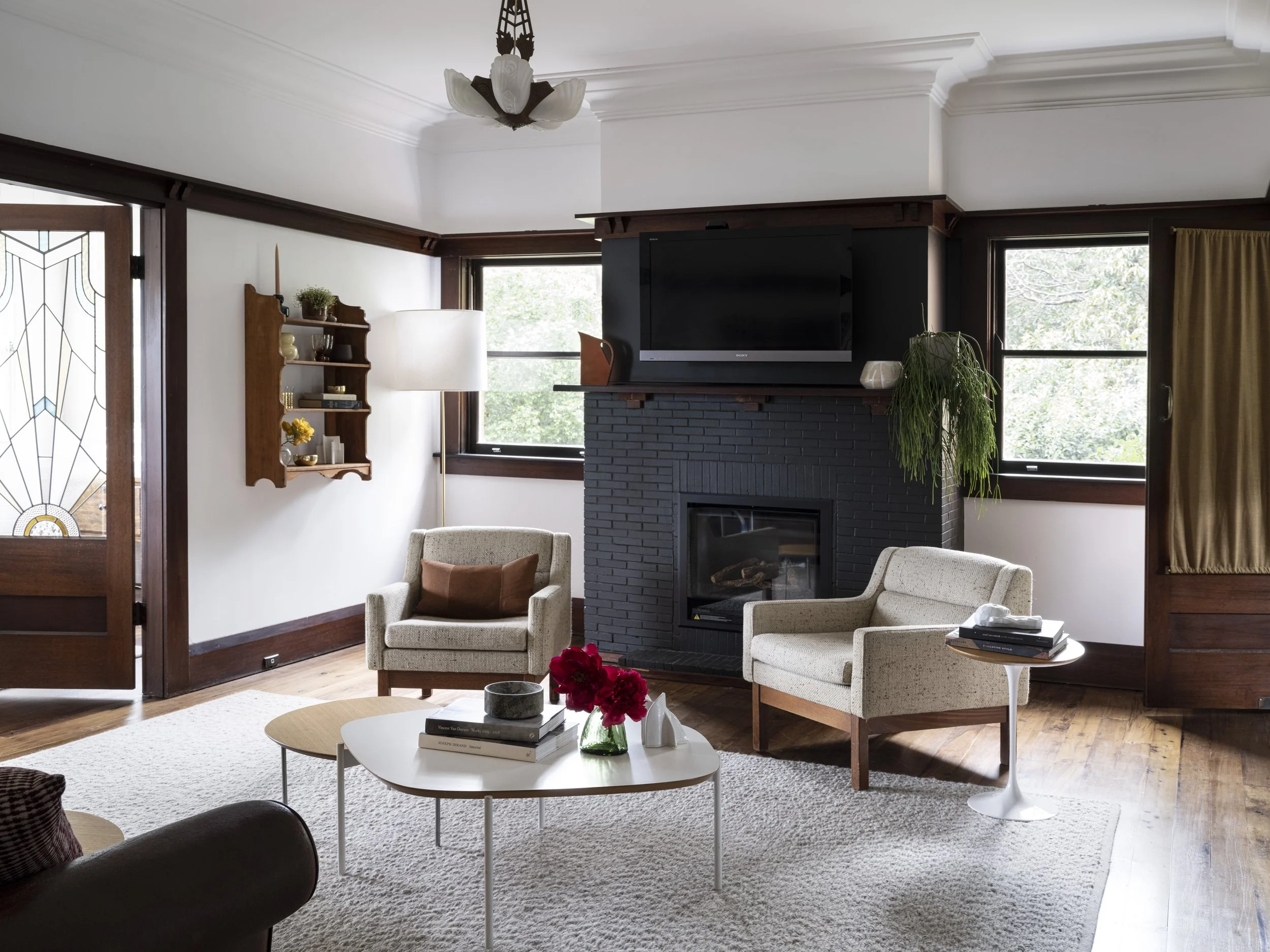 Living room with two white armchairs, a white coffee table with books and flowers, a fireplace with a TV on the mantel, windows with wood trim, and a cream-colored rug.