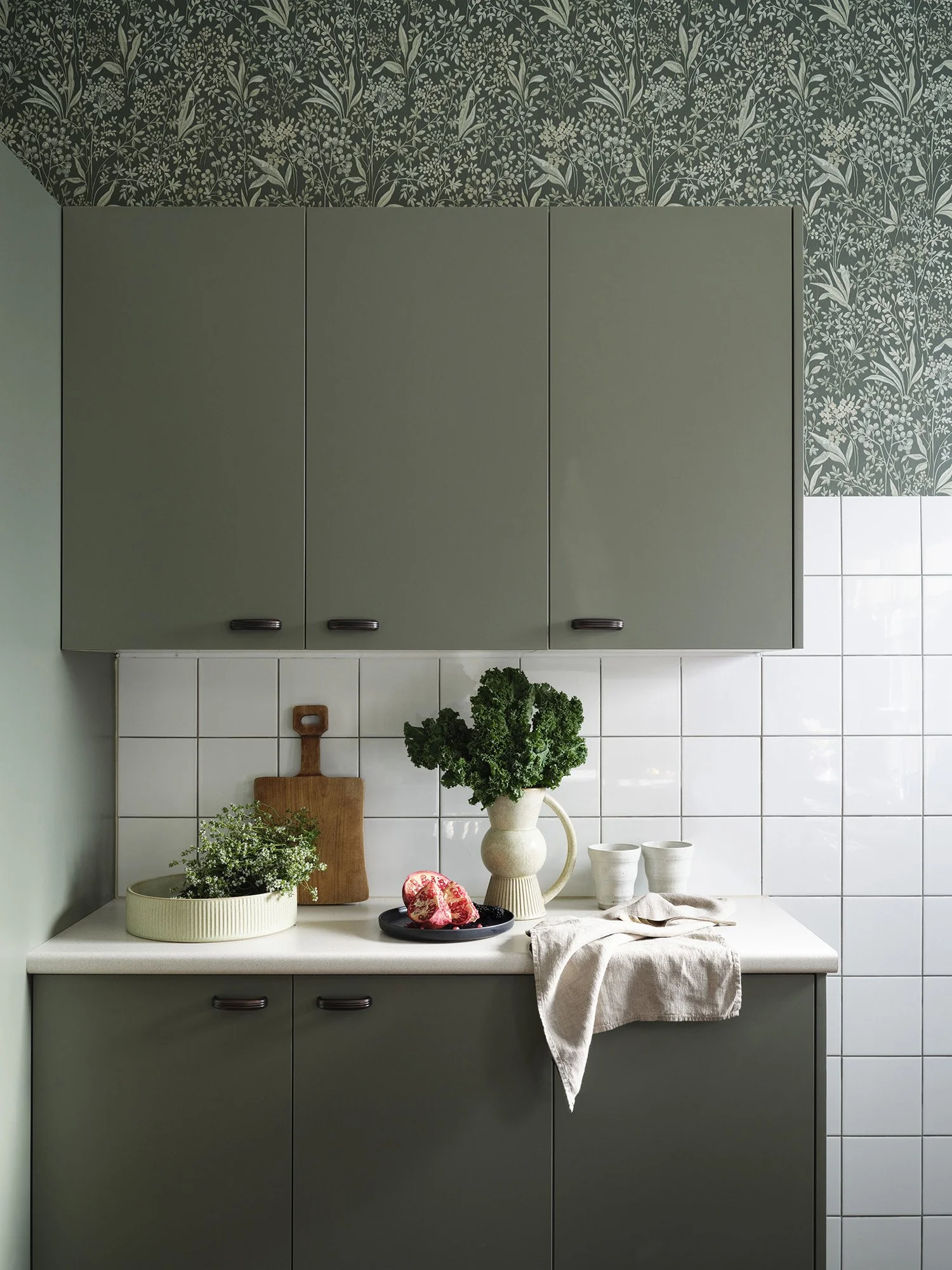 Kitchen with green cabinets, white tiled wall, and countertop decorated with plants, a wooden cutting board, and dishes.