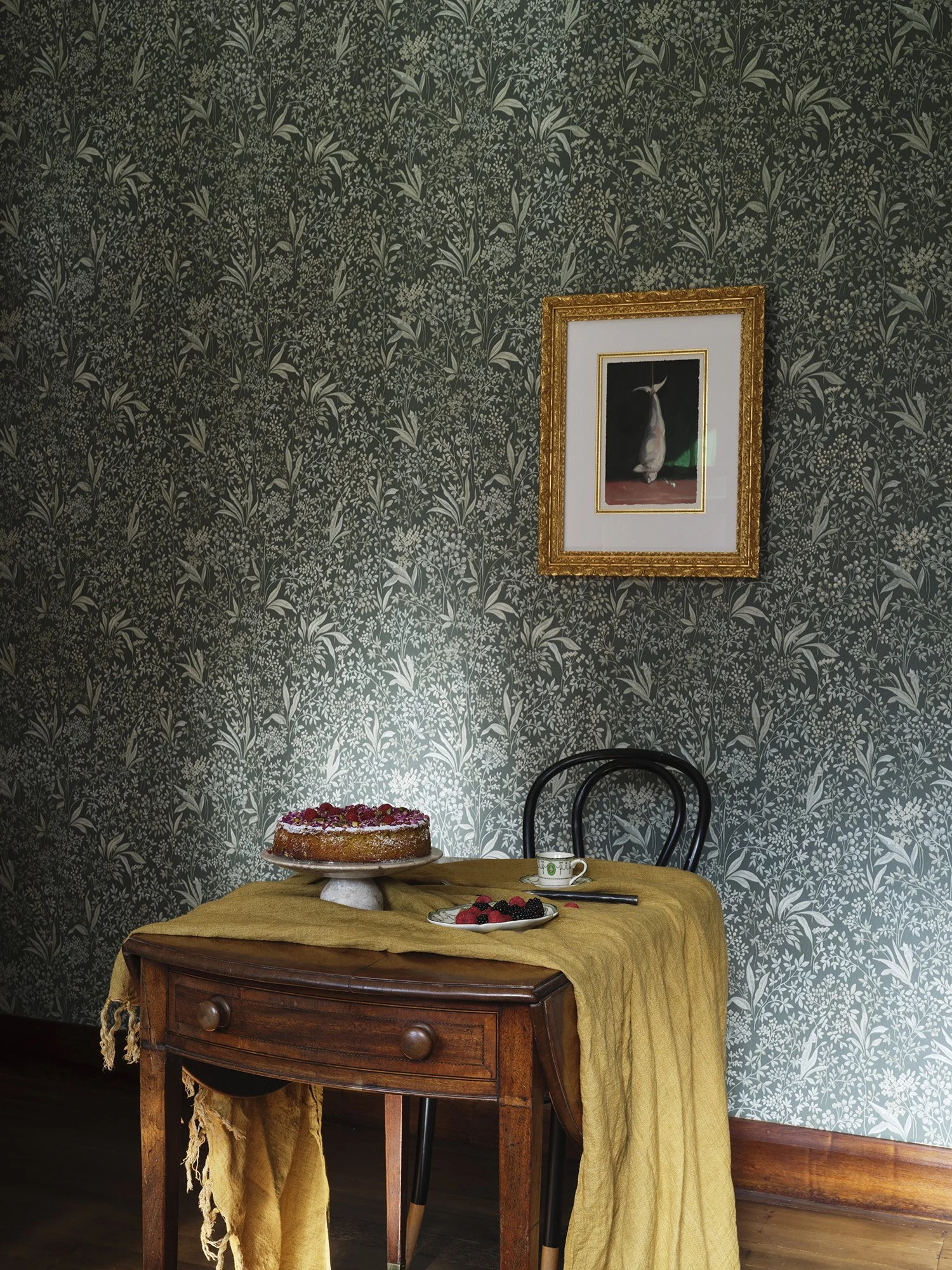 A wooden table with a yellow cloth, topped with a cake, a plate of berries, a spoon, and a teacup. There are two black chairs behind the table. The background features green wallpaper with white floral patterns and a framed painting of a hanging fish.