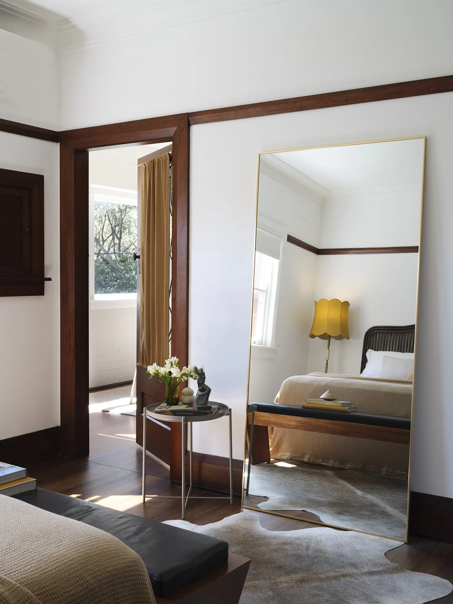 A bedroom with a large mirror, a bed with a beige blanket, a yellow lamp, a small side table with flowers and decorative items, visible windows with natural light.