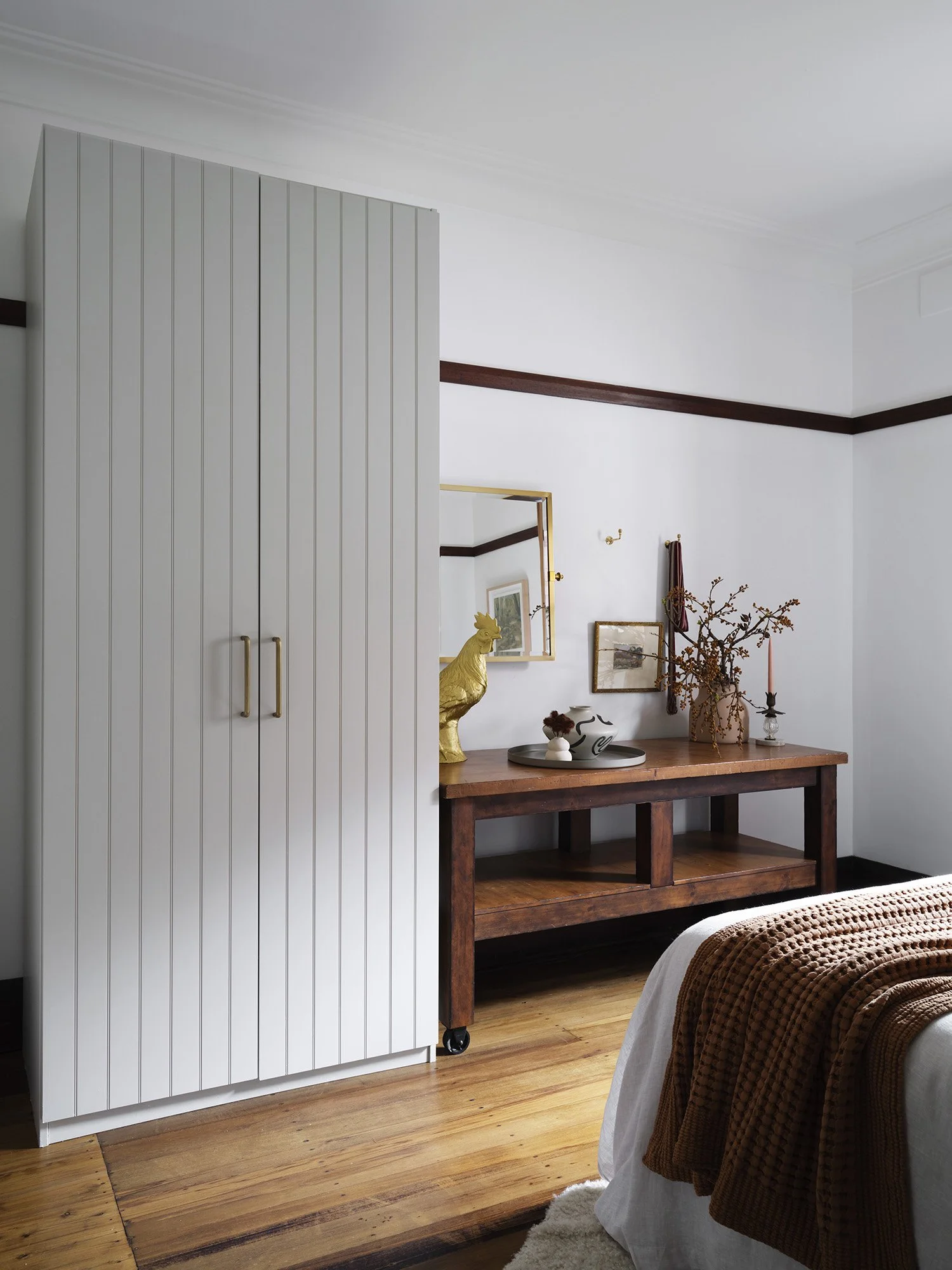 A bedroom with a white vertical-paneled wardrobe, a wooden console table with decorative items, a mirror, picture frames, a vase with dried branches, a candle, and a bed with a brown blanket.