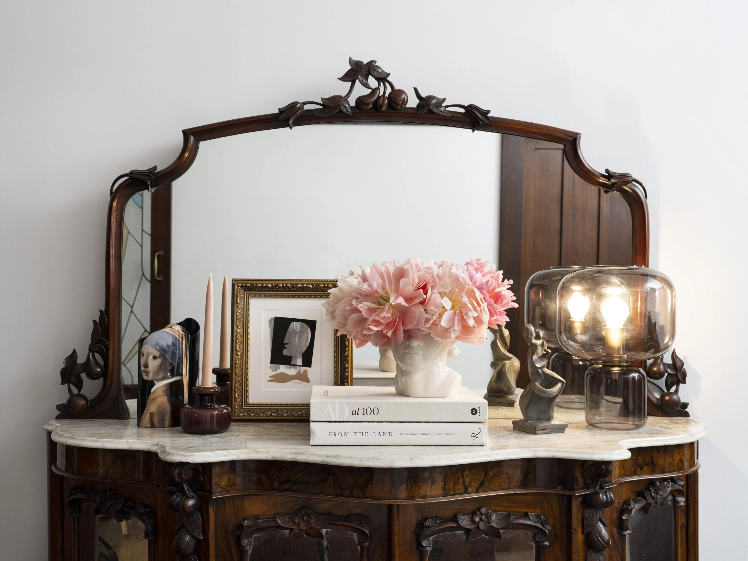 A vintage wooden dresser with a large mirror, decorated with a floral arrangement, framed photo, books, candles, and sculpture pieces.
