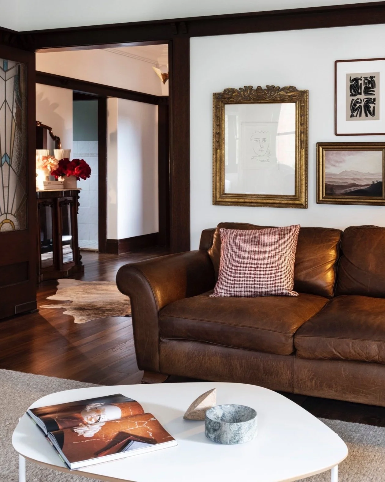 Living room with a brown leather sofa, a white coffee table with an open magazine, and four framed pictures on the wall, including a framed line drawing and landscape painting, with a hallway visible in the background.