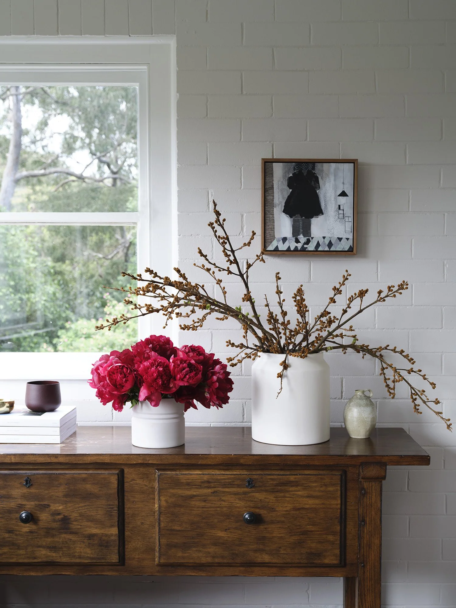 A wooden sideboard with a white vase holding pink peonies and a white vase with branches, set against a white brick wall with a window and a black and white artwork.