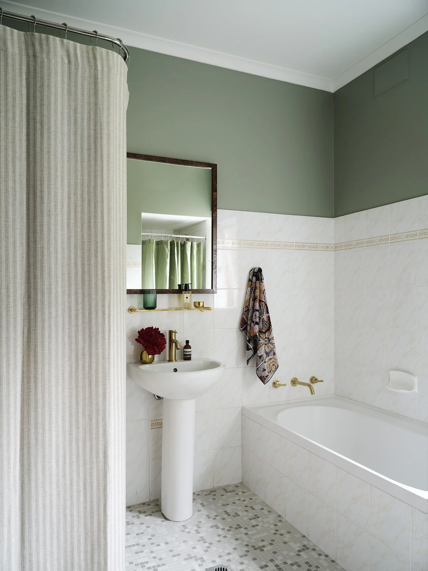 A small bathroom with a green upper wall, white tiled lower wall, and a mosaic tile floor. Features include a small pedestal sink with a gold faucet and a mirror, a bathtub with a gold faucet, a wall-mounted towel rack with a patterned towel, and a shower curtain on a curved rod.