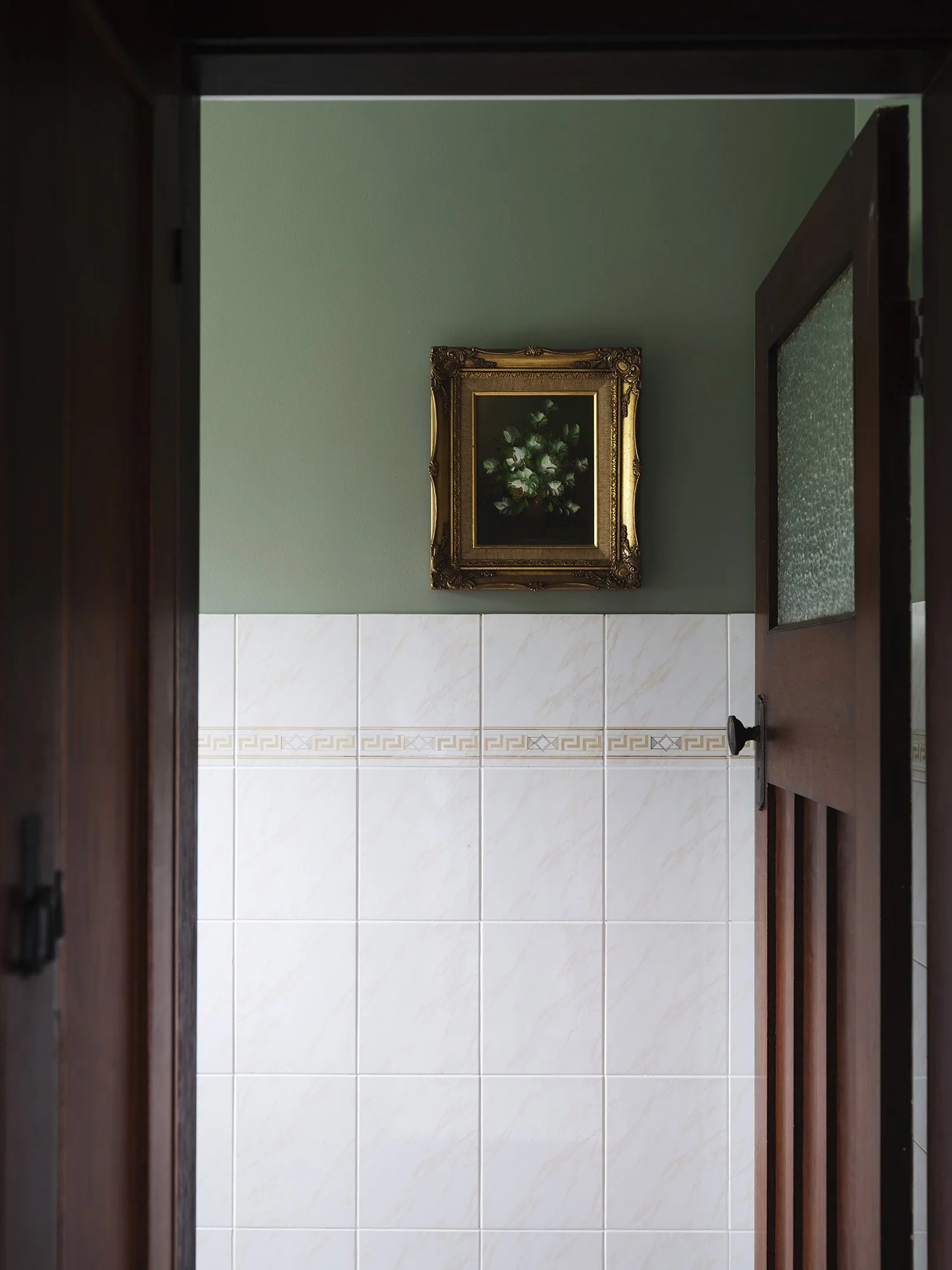 Looking through a doorway into a bathroom with white tiles, a green wall, a framed painting of white flowers, and a wooden door.