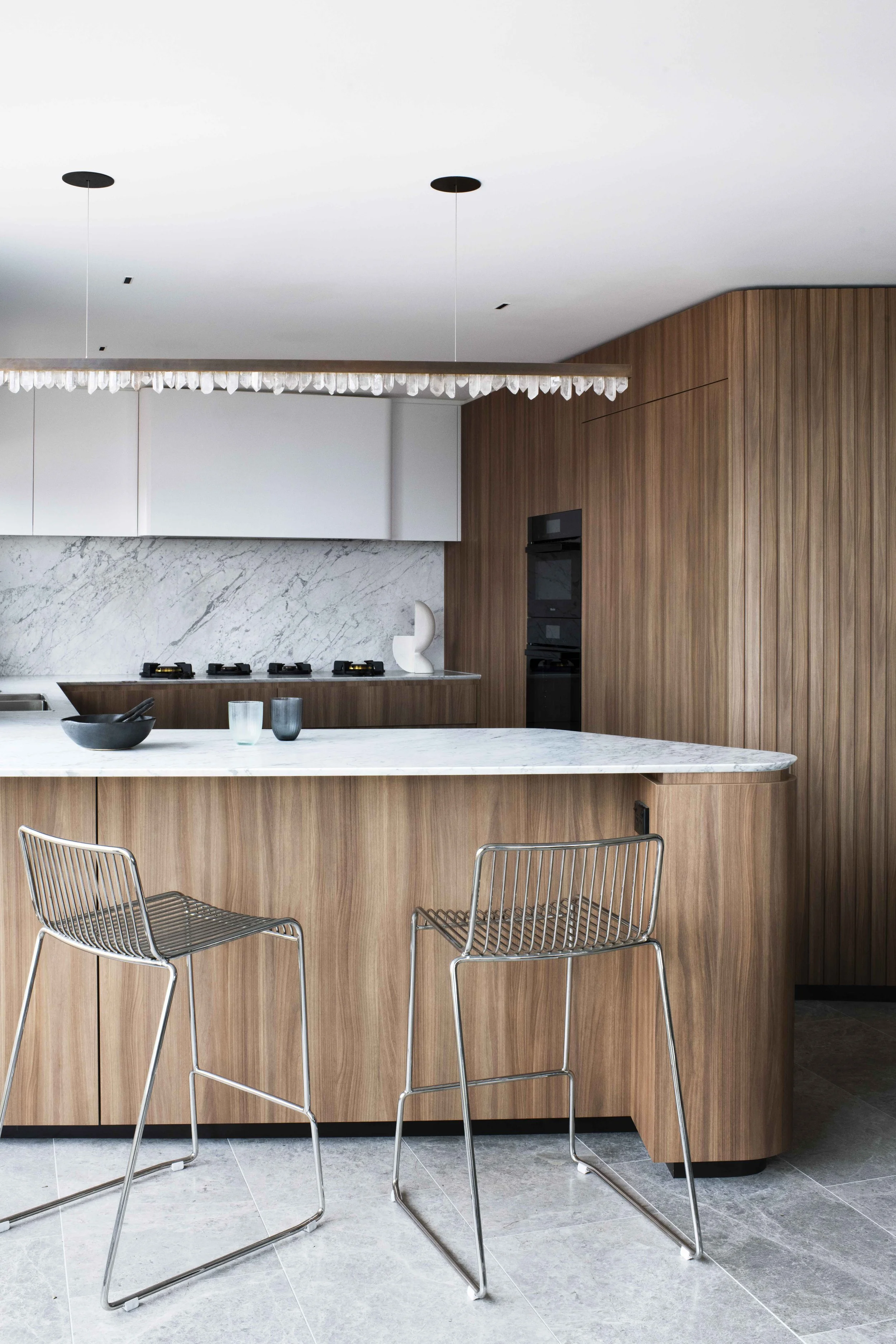 Modern kitchen with marble island, wooden cabinetry, and minimalistic decor, including two metal barstools, black bowls, and glass cups.