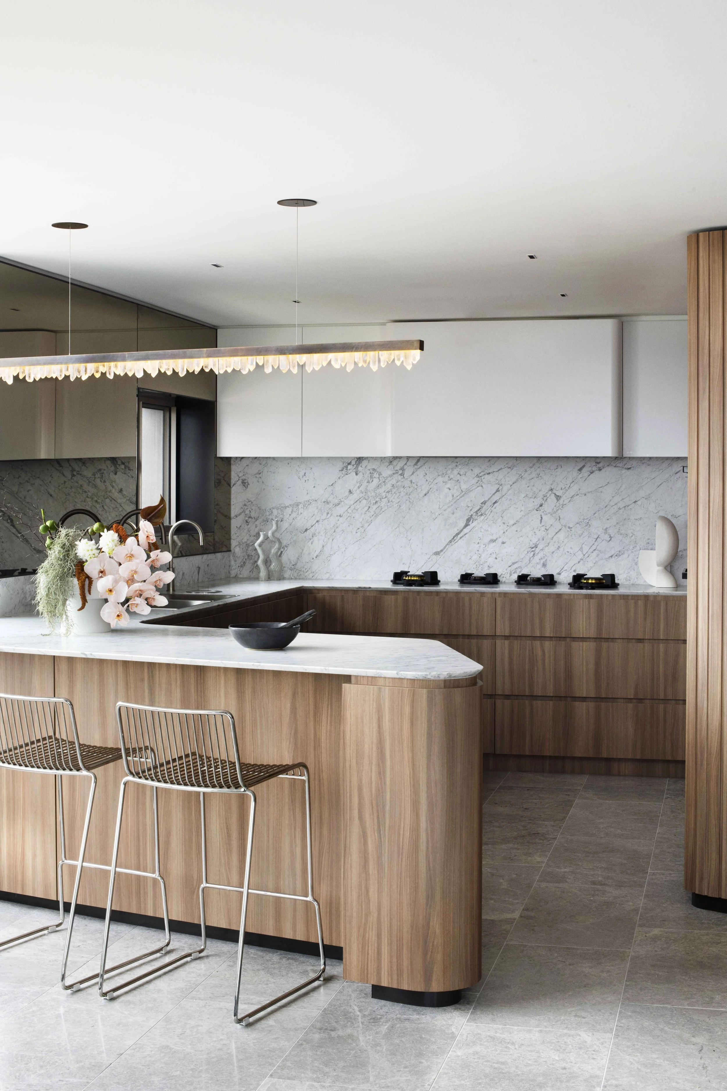 Modern kitchen with wooden cabinetry, white marble countertops, a flower arrangement, and a decorative sculpture, with two wire bar stools.