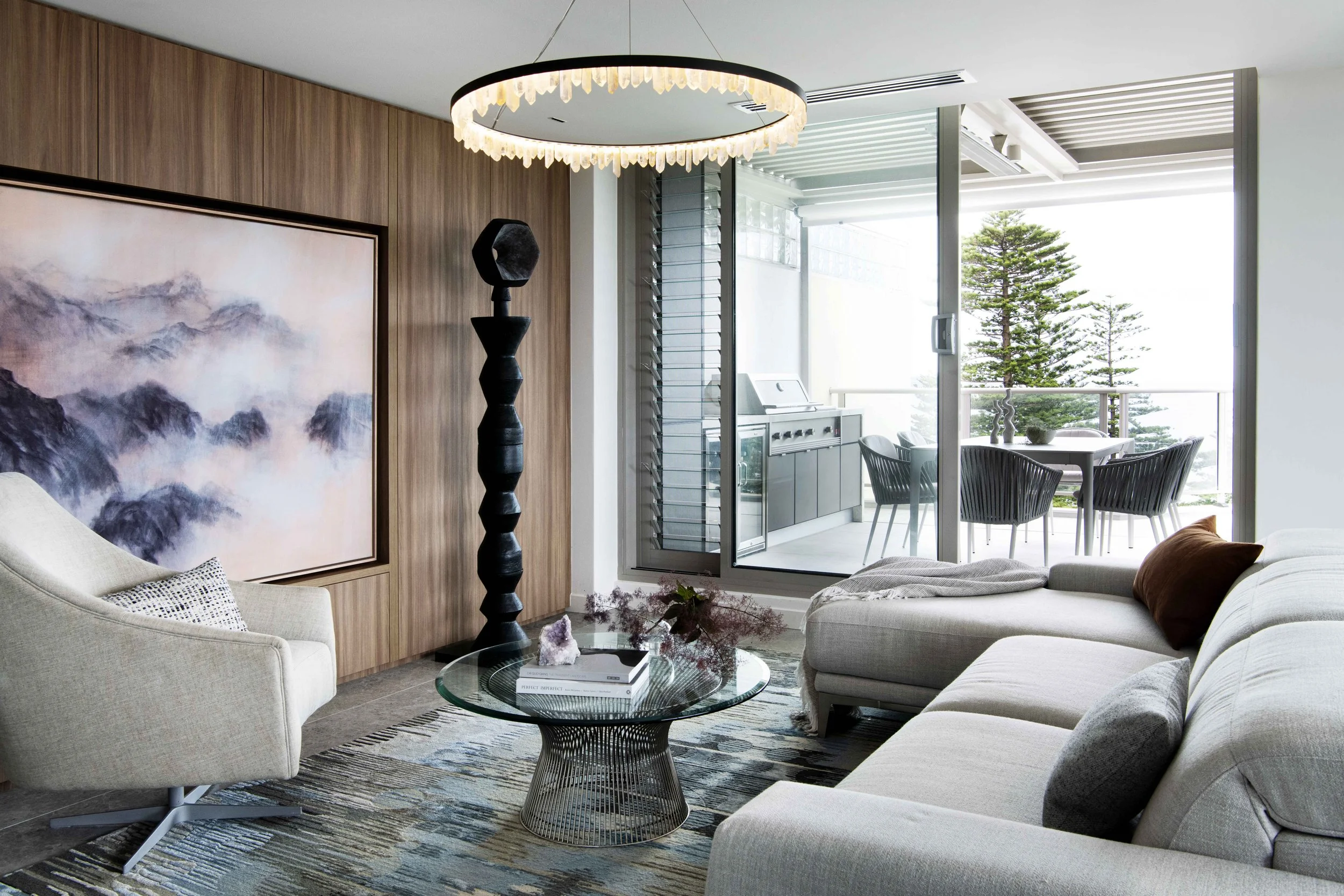 Modern living room with a beige sofa, armchair, glass coffee table, wooden wall panel, large painting, black decorative sculpture, and a balcony with outdoor furniture and trees in the background.