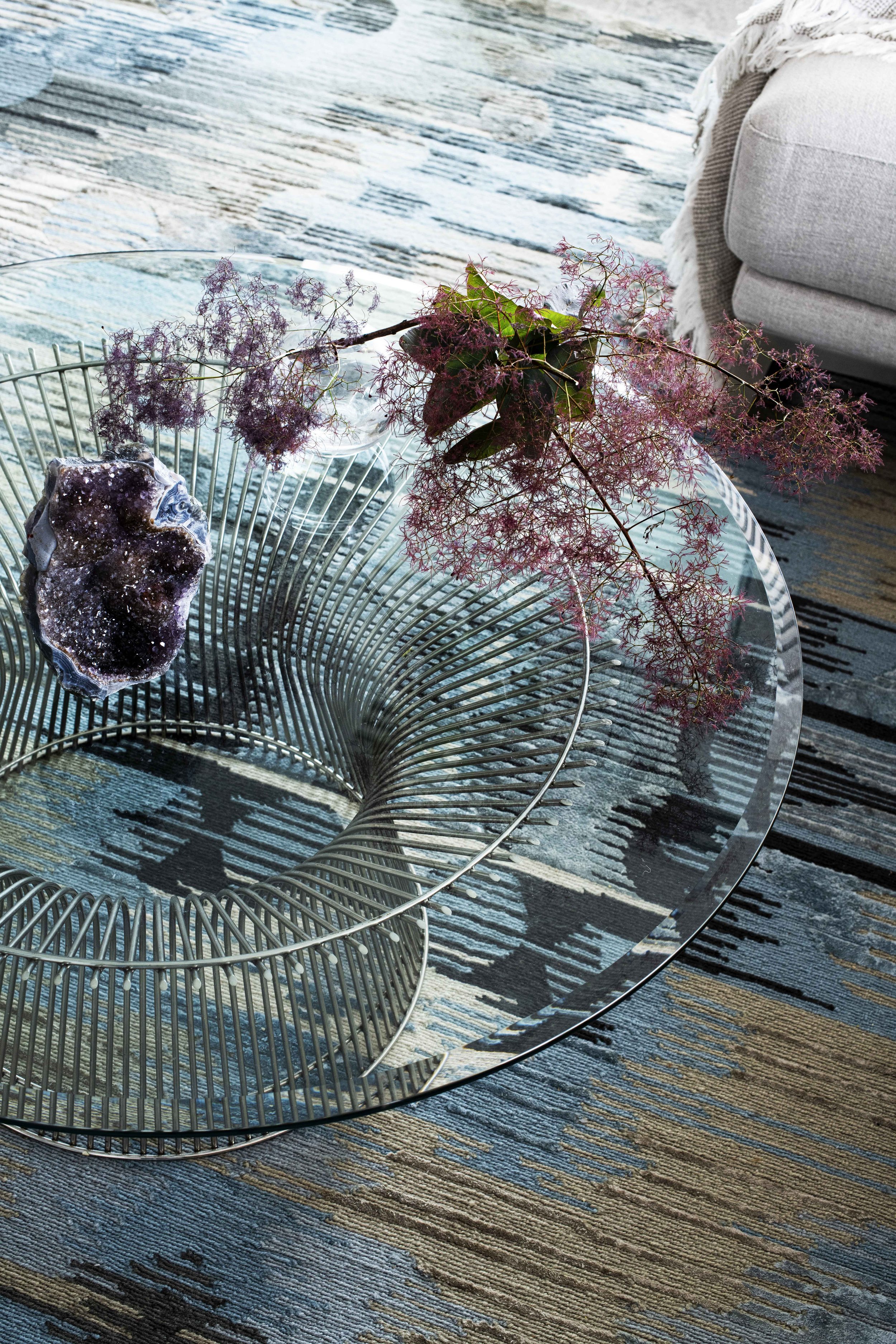 A round glass coffee table with a wireframe metal base, decorated with a purple, black, and white geode and pink flowers in a glass vase. The table is on a colorful patterned rug in a living room with a gray couch visible in the background.