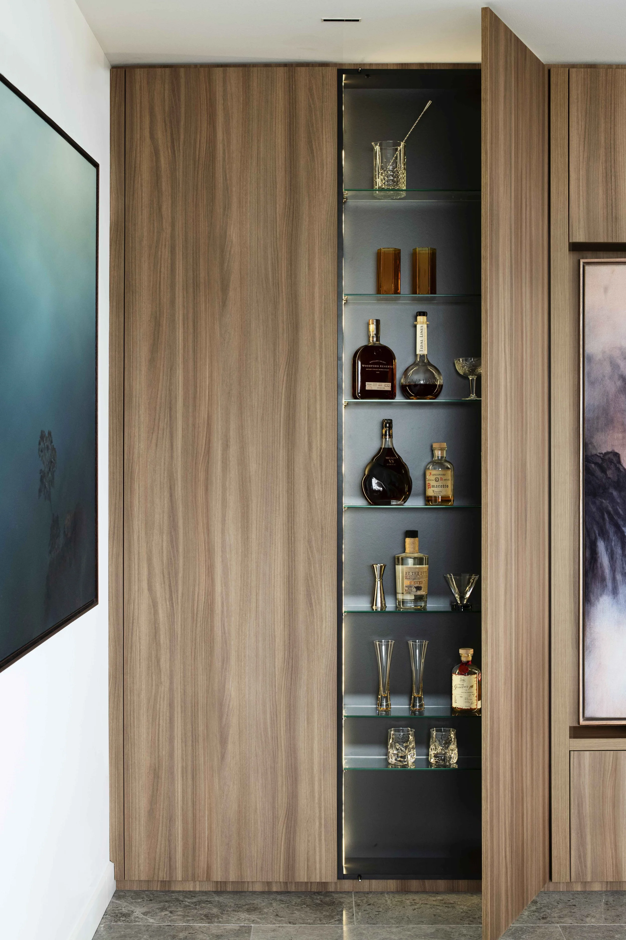 Built-in wooden cabinet with glass shelves holding liquor bottles and glasses, partially open door revealing the display.