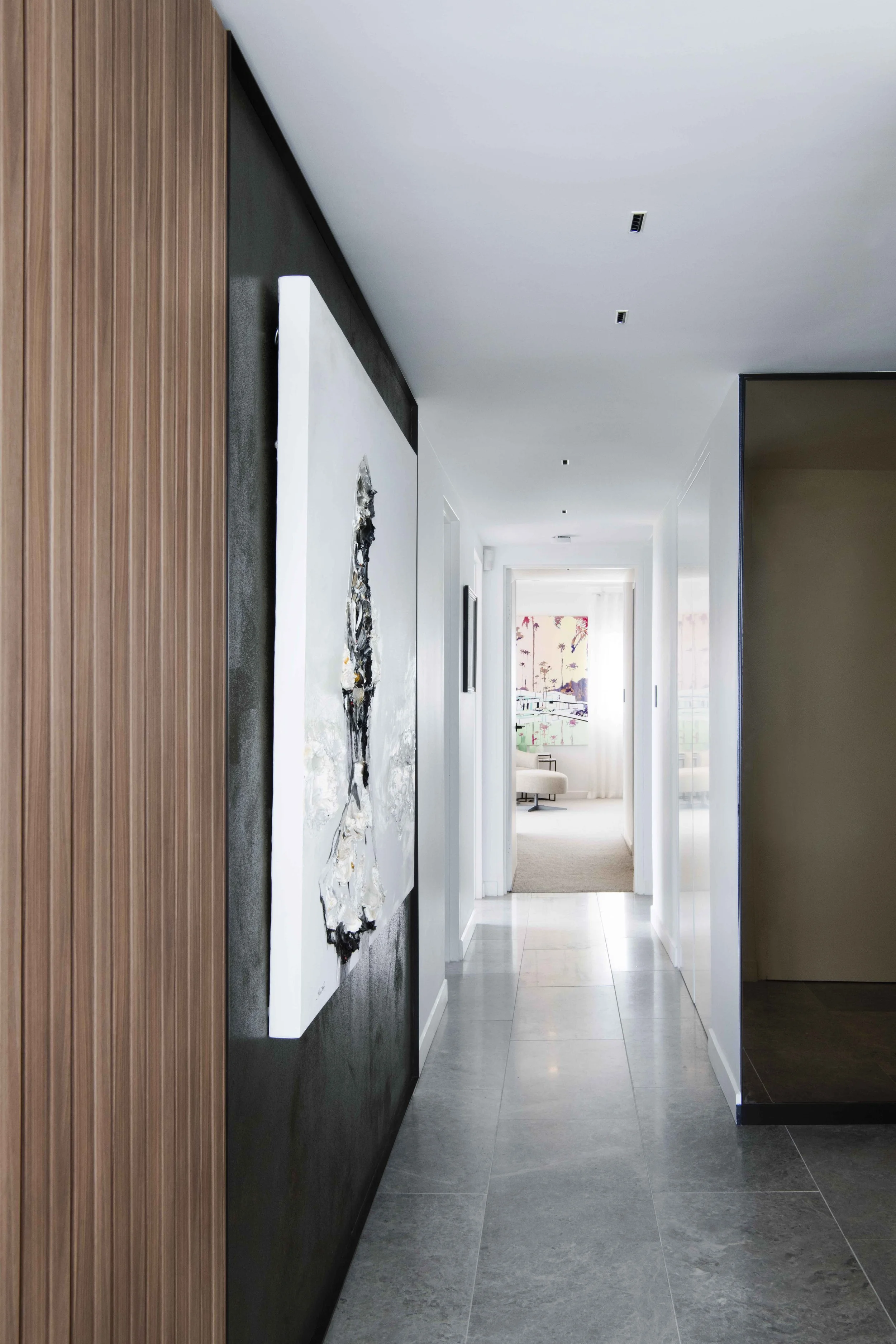 Modern hallway with gray tiled floor, white walls, and a black accent wall on the left with a large abstract painting. Natural light coming from a room at the end of the corridor, which contains a small table and artwork with palm trees.