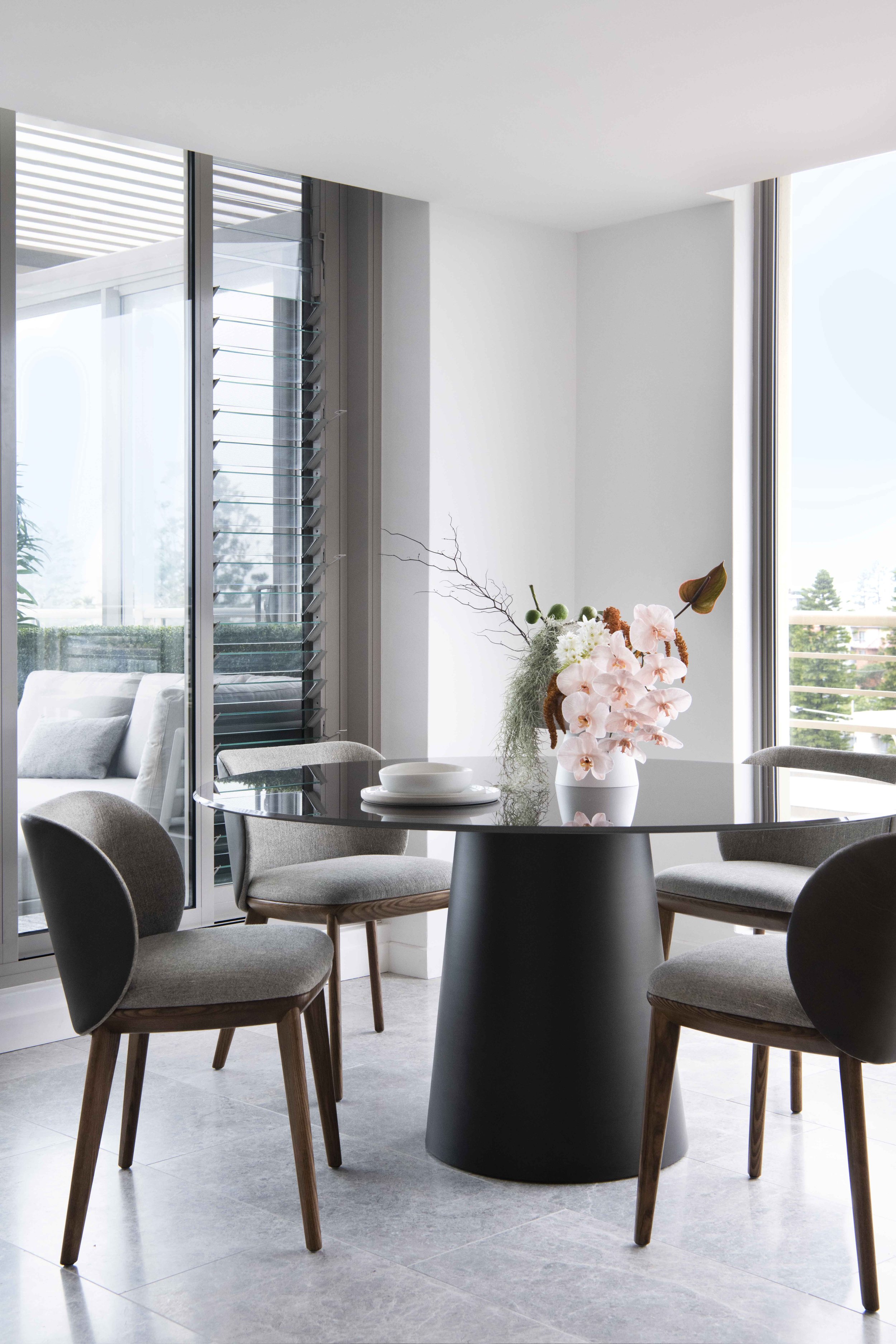 Modern dining room with a round black table, four cushioned chairs, and a floral centerpiece, near large windows with view of balcony and outdoor greenery.