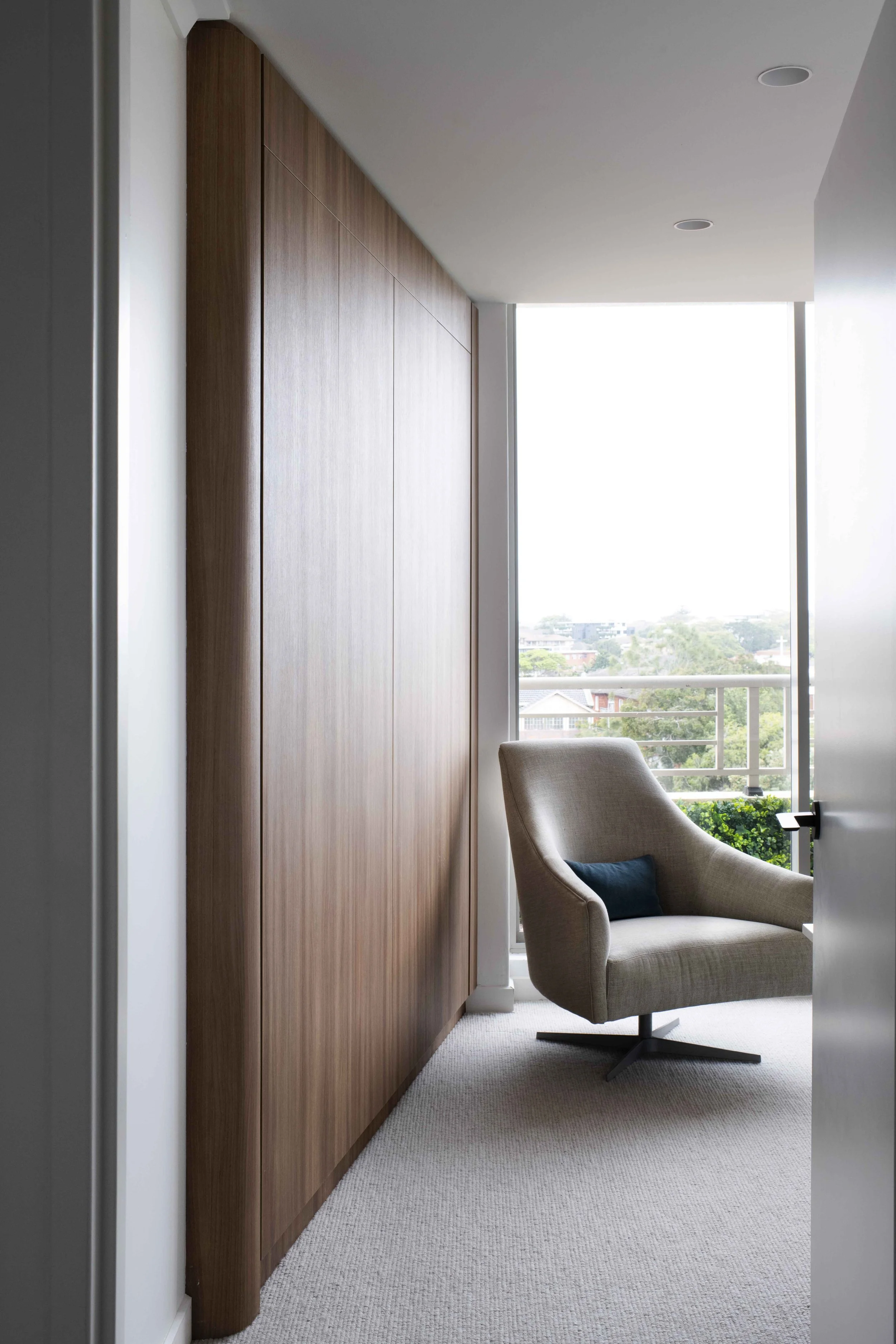 A corner of a room with a large wooden cabinet, a beige armchair with a dark cushion, and a large window with a view of trees and buildings outside.