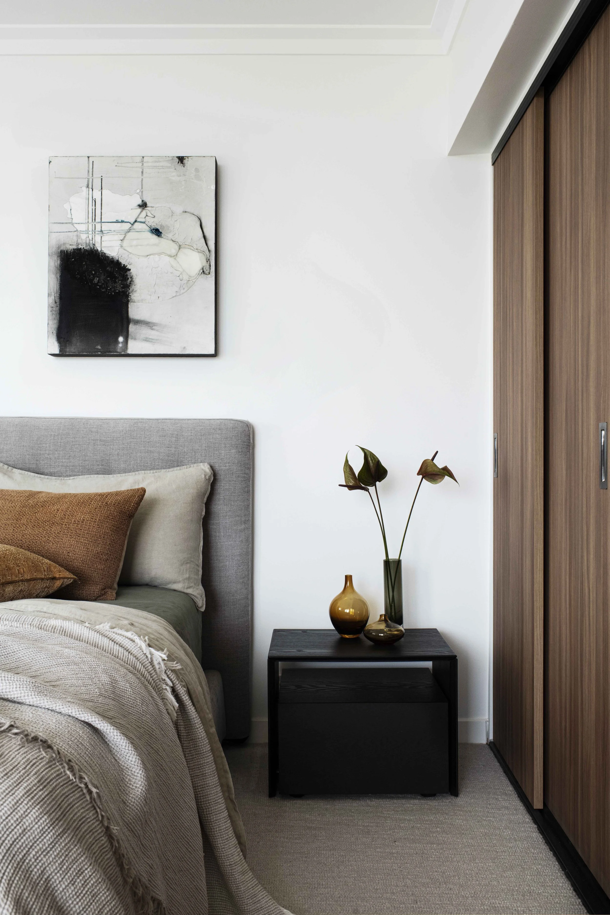 A bedroom corner featuring a bed with neutral-toned bedding and pillows, a small black nightstand with three decorative vases, a white wall with a black and white abstract painting, and a wooden closet with sliding doors.