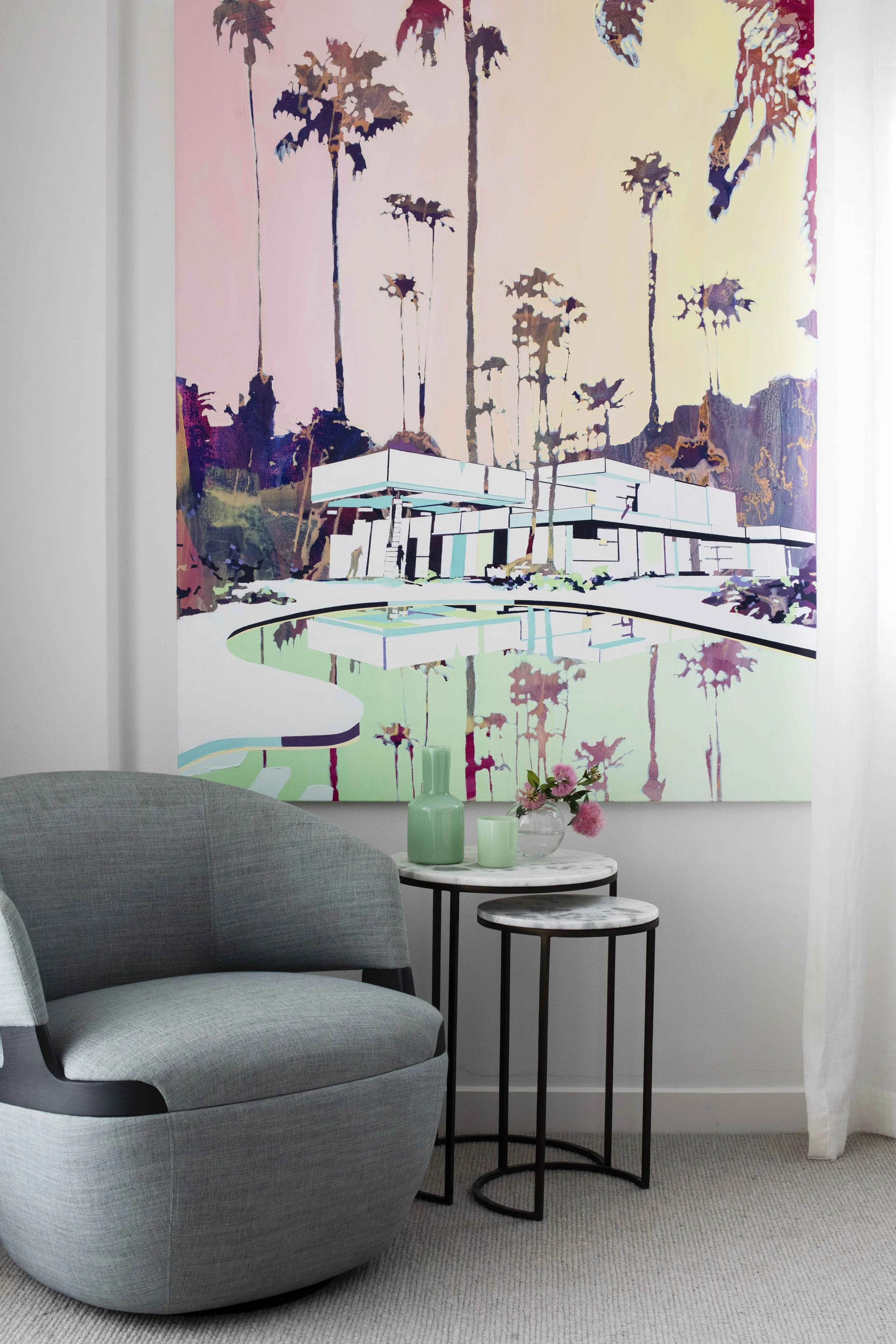 Interior living room with a large colorful painting of a modern house and palm trees on the wall, a gray armchair, and two small round tables with green vases and pink flowers.