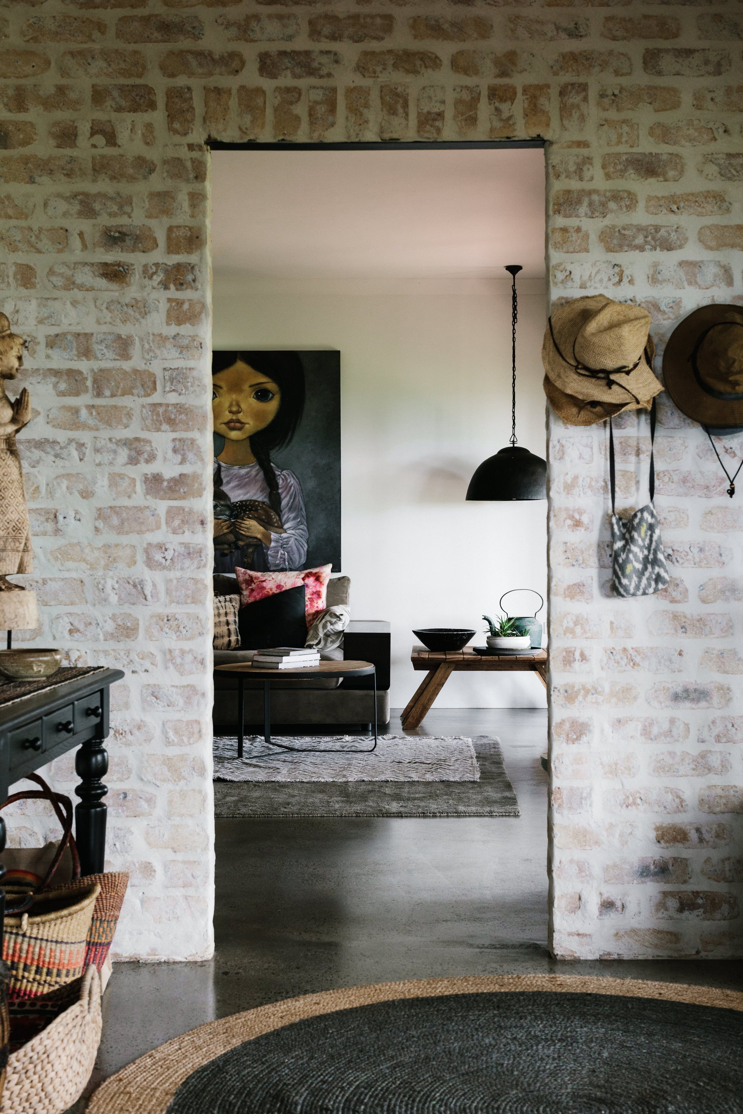 Living room viewed through a brick archway. Features a large painting of a girl holding a dog, a gray sofa with colorful cushions, a wooden bench, a black pendant light, and a wall with hats and bags hanging.