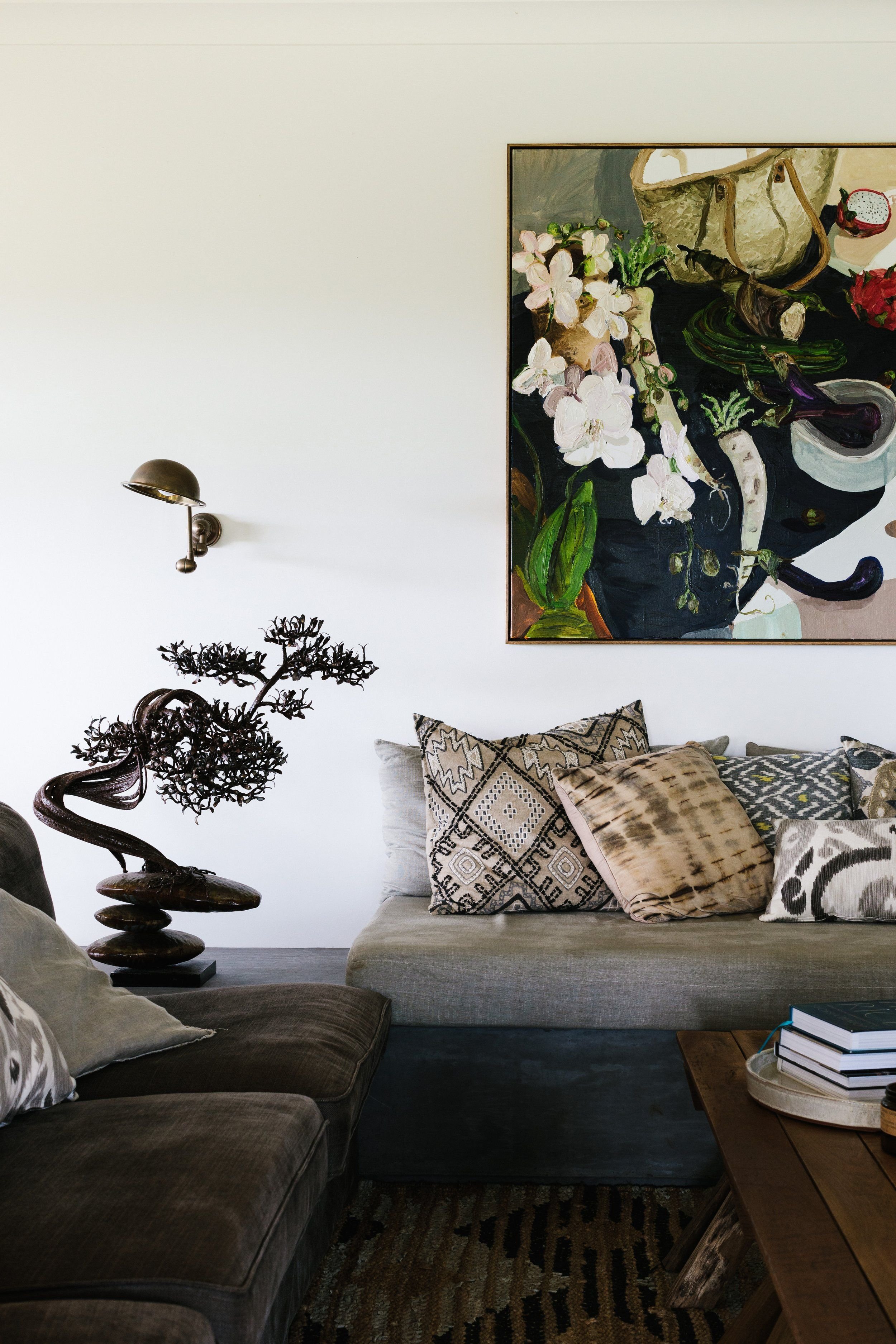 Living room with beige and brown furniture, patterned cushions, a wooden side table with books, a decorative sculpture of a tree, a modern wall lamp, and a colorful still life painting of flowers and objects.