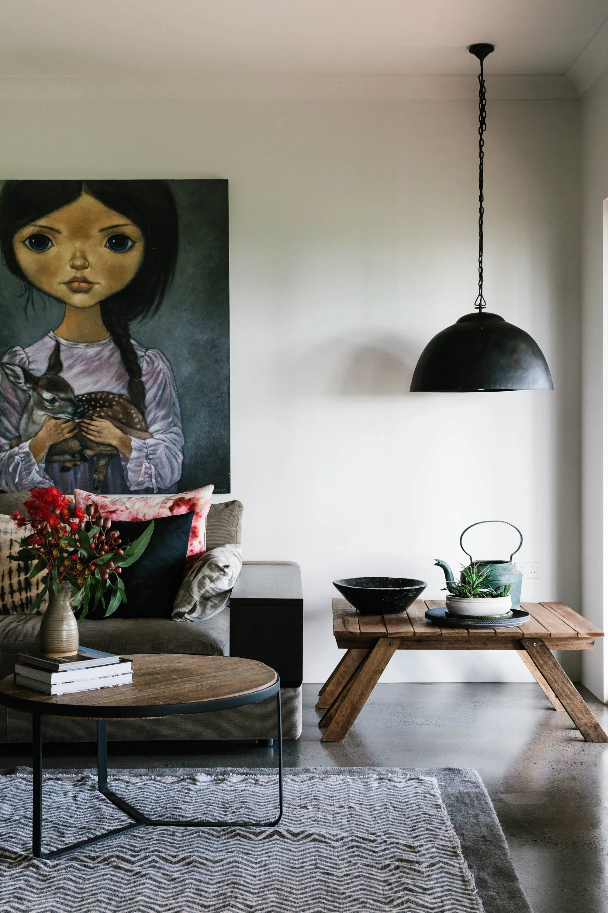 Modern living room with a sofa, a coffee table, and a wooden console table. A large painting of a girl holding a deer hangs on the wall. There is a black pendant light fixture hanging from the ceiling, and decorative items such as a teapot, bowl, and plants on the tables. A vase with red flowers sits on the coffee table, and a patterned rug covers part of the floor.