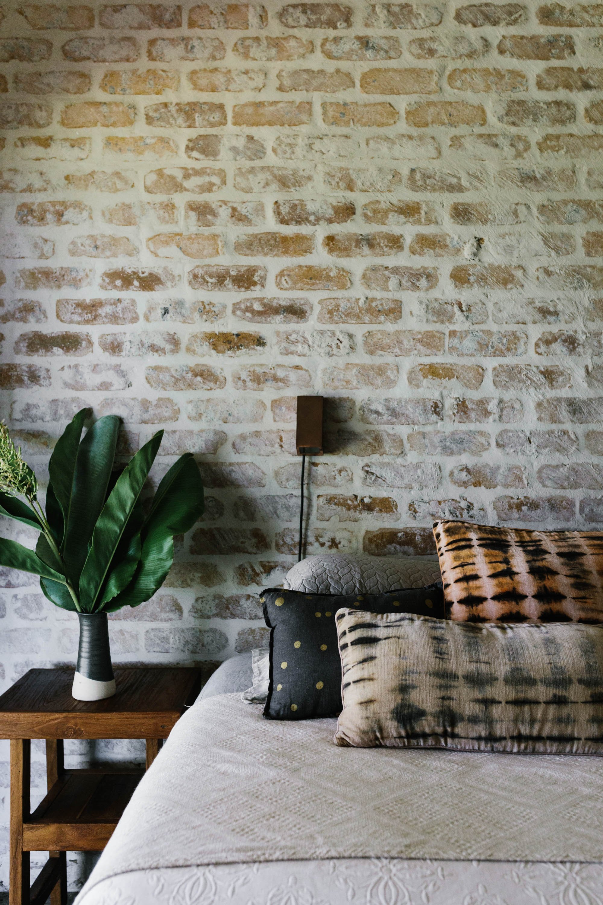 A bedroom with a brick wall, a plant on a wooden table, and pillows on a bed with a beige bedspread.