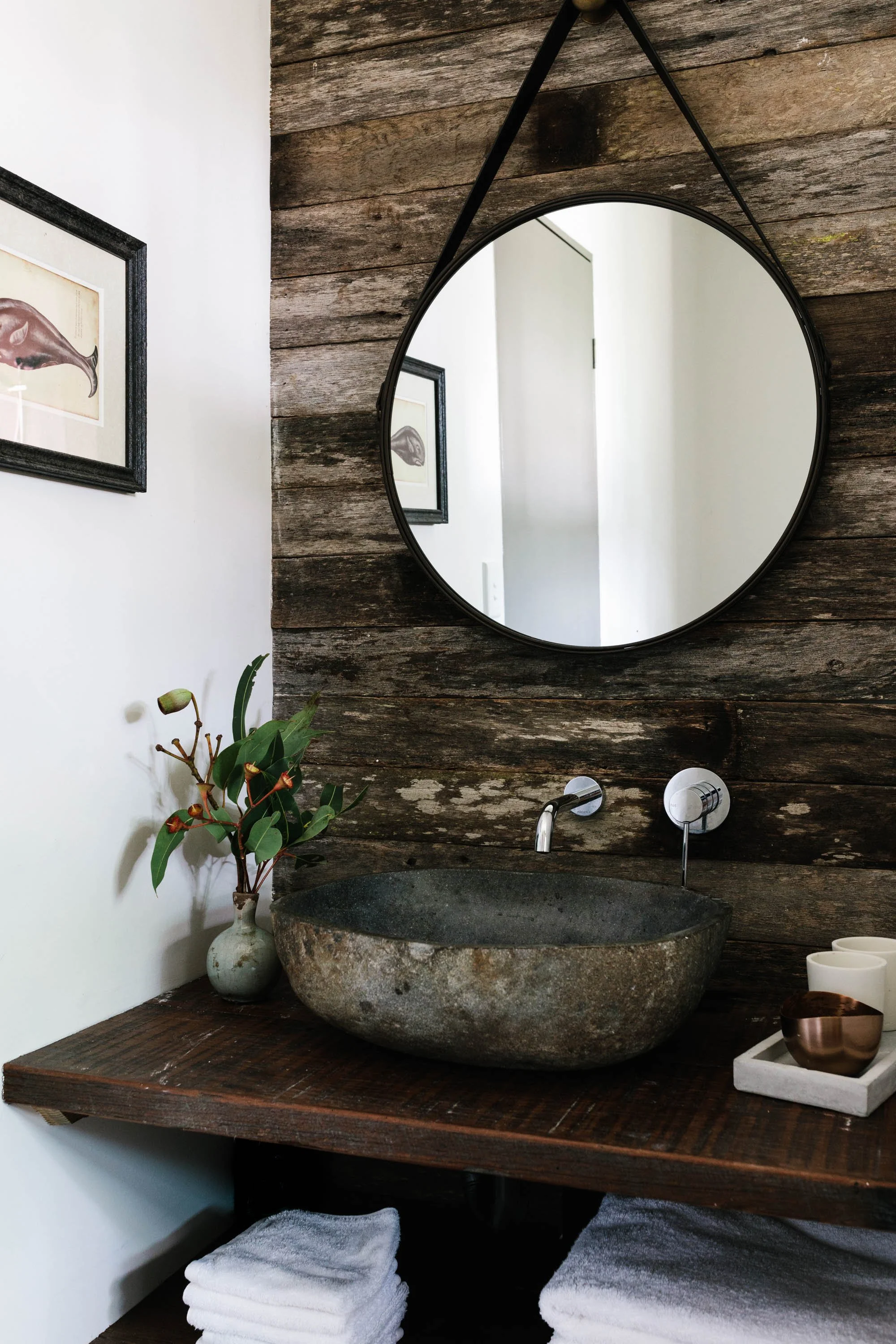 Rustic bathroom with a stone sink, round mirror, wooden countertop with folded towels, and framed artwork of whales on the wall.