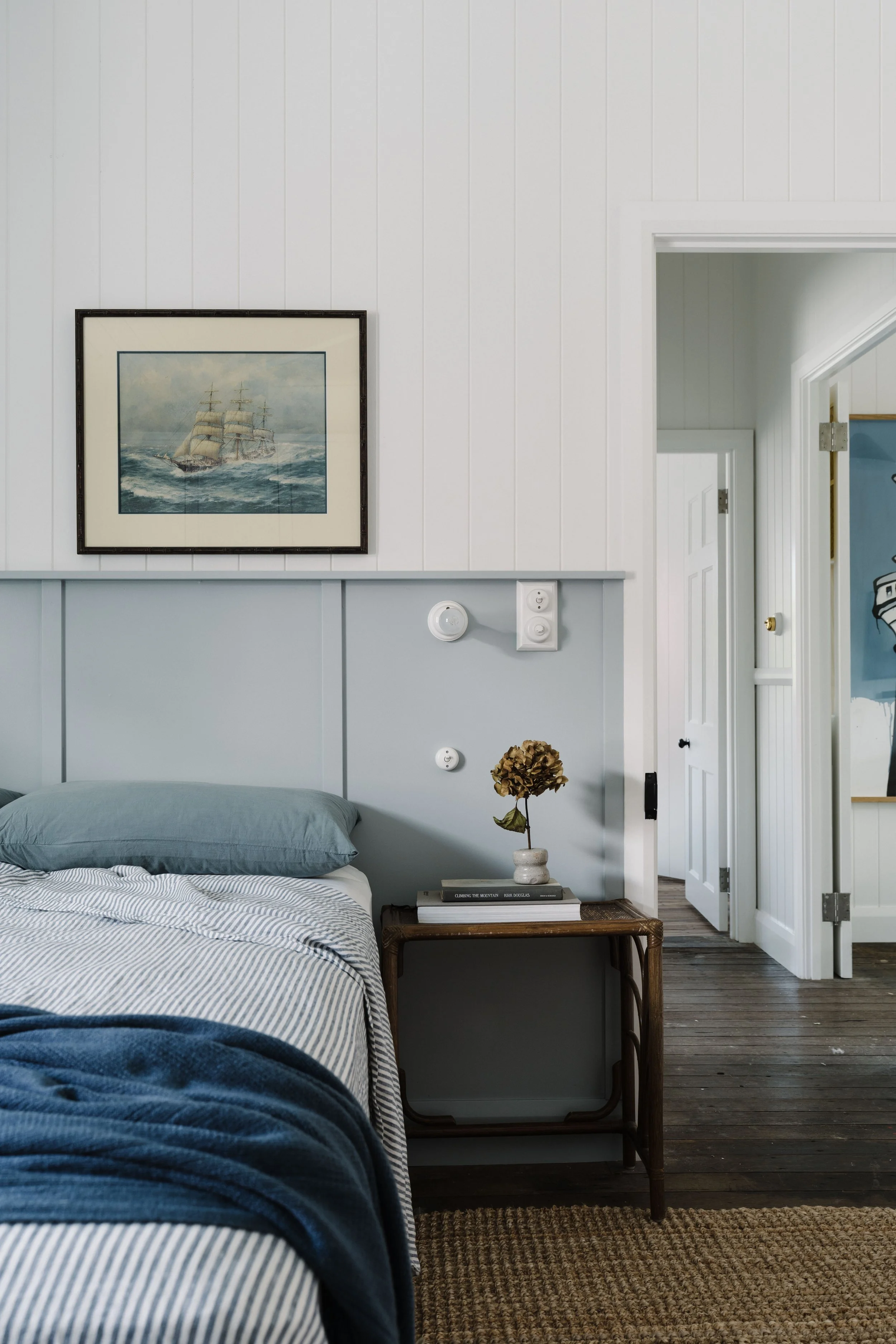 A bedroom with light blue walls, a bed with a striped comforter, a wooden nightstand with books and a dried flower in a vase, a framed painting of a sailing ship, and a doorway leading to another room with wooden floors.