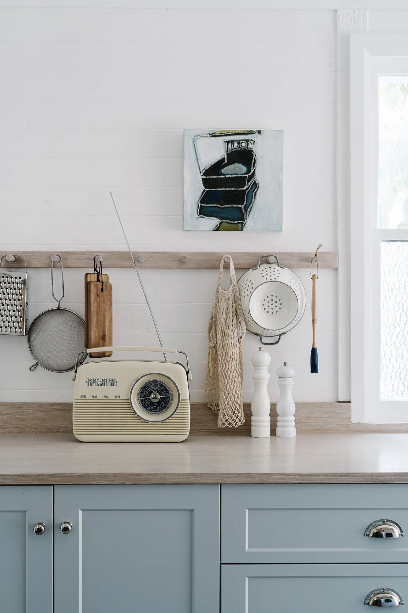 Vintage radio on kitchen counter with kitchen utensils hanging above, including a cutting board and a strainer, in a white kitchen with blue cabinets and a window.