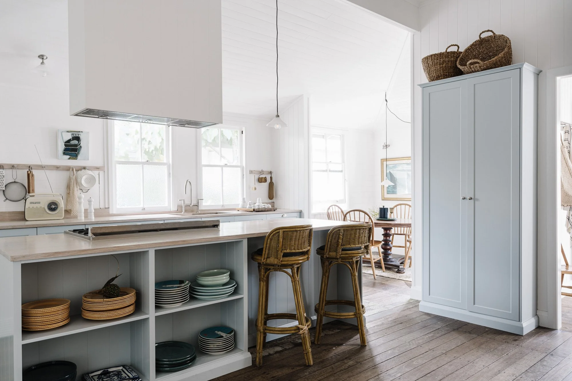 Bright, airy kitchen with white walls and wooden flooring, featuring a light blue cabinet, wicker bar stools, and an open shelving unit with dishes. Sunlight streams through multiple windows.