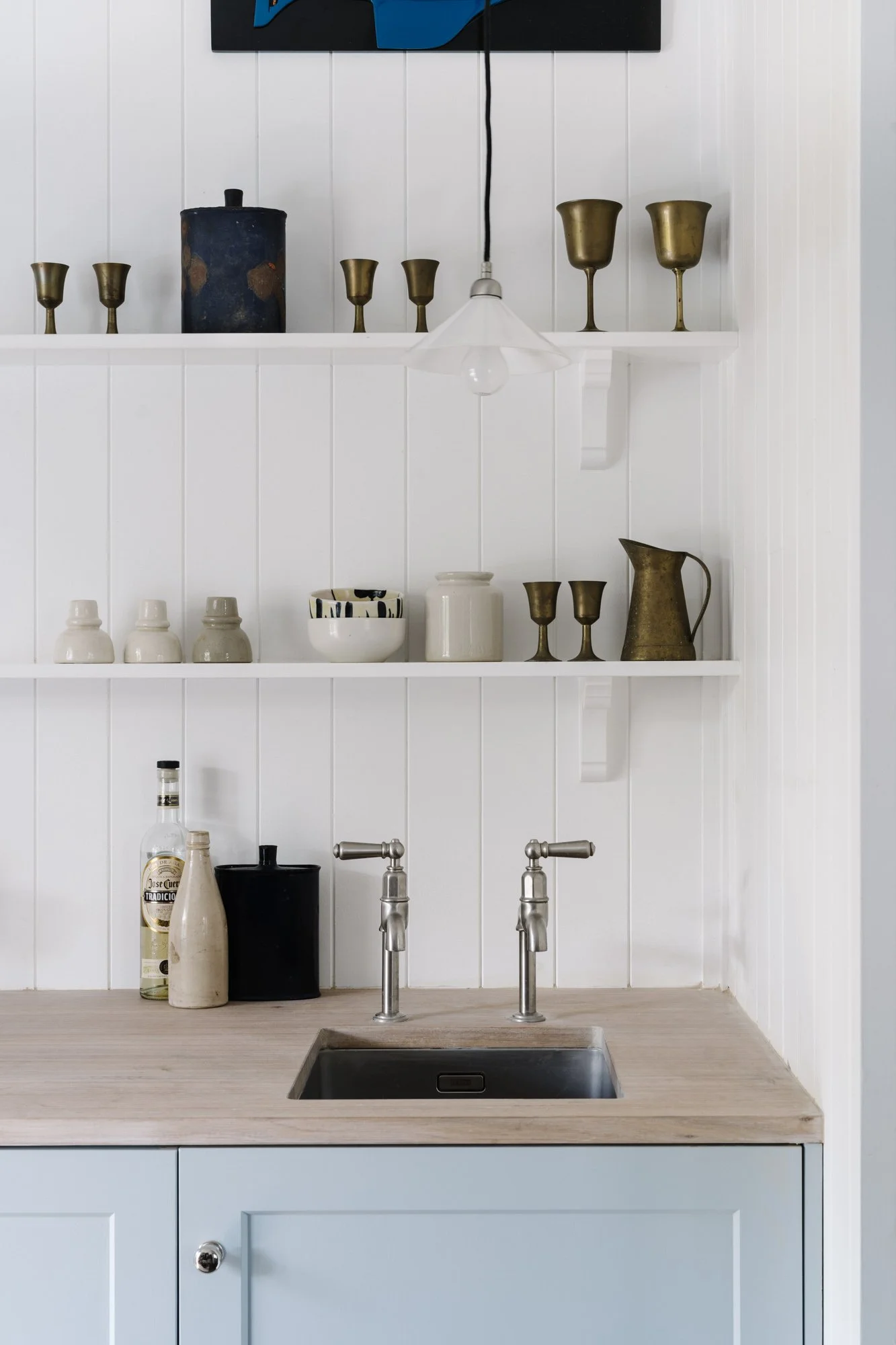 A kitchen with white vertical panel walls, open shelves holding various cups, bowls, and pitchers, a gray countertop with a stainless steel sink, and a light blue cabinet below the counter.