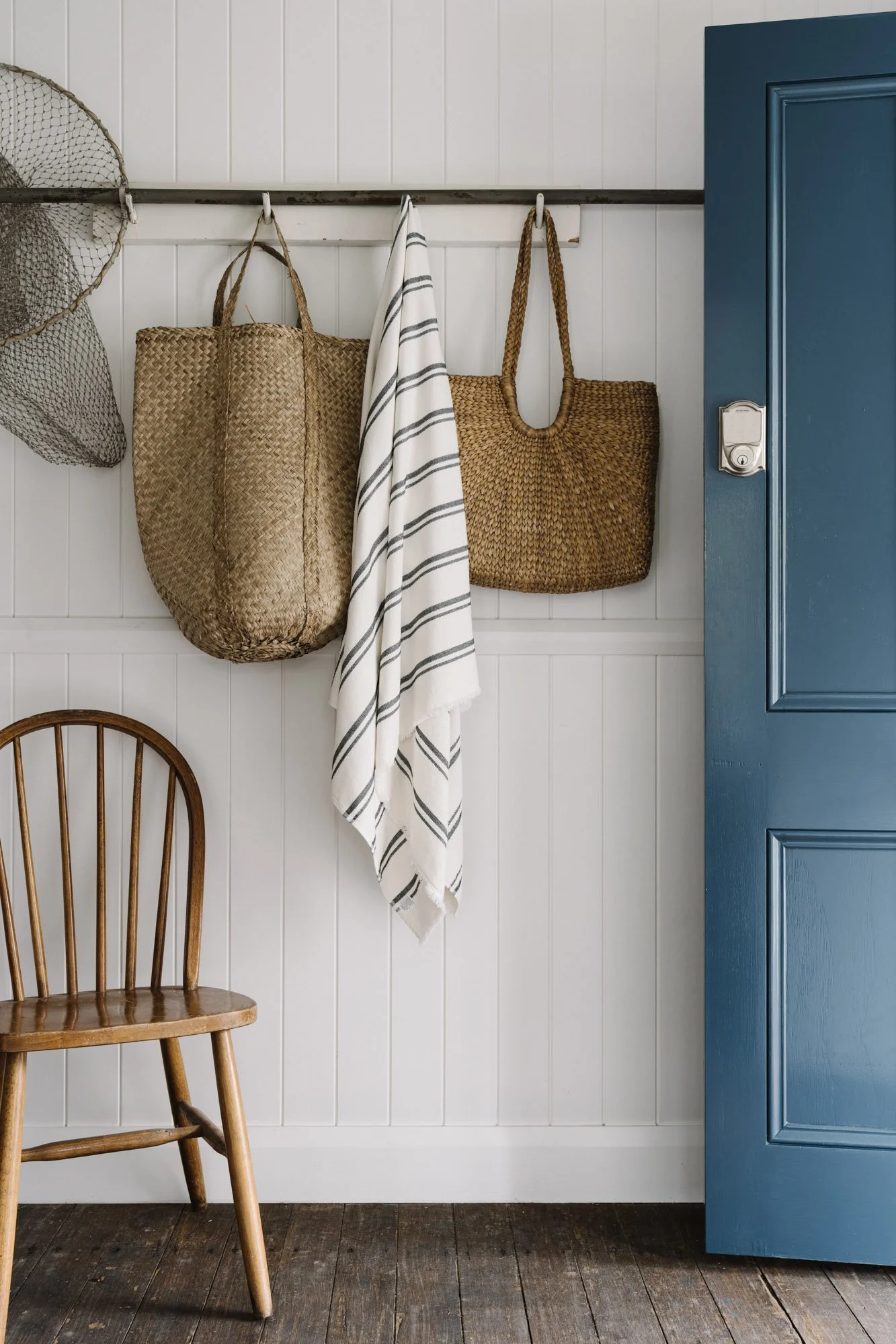 Coastal entryway with hanging woven bags, a striped cloth, a wooden chair, a blue door, and white paneled wall.
