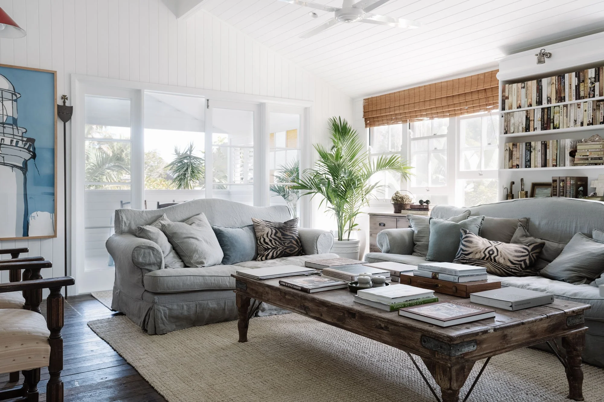 Bright living room with white walls, large windows, and a wooden coffee table surrounded by light gray sofas with various cushions. A large potted plant and bookshelves are visible in the background.