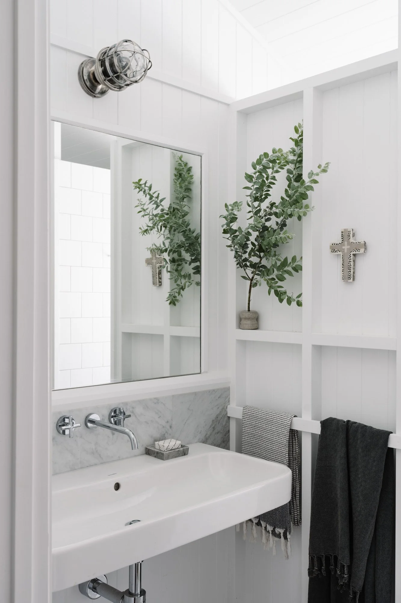 A small white bathroom with a rectangular vessel sink, wall-mounted faucet, mirror, and a wall decorated with a green plant in a gray pot and crosses, with black and gray towels hanging on the right side.