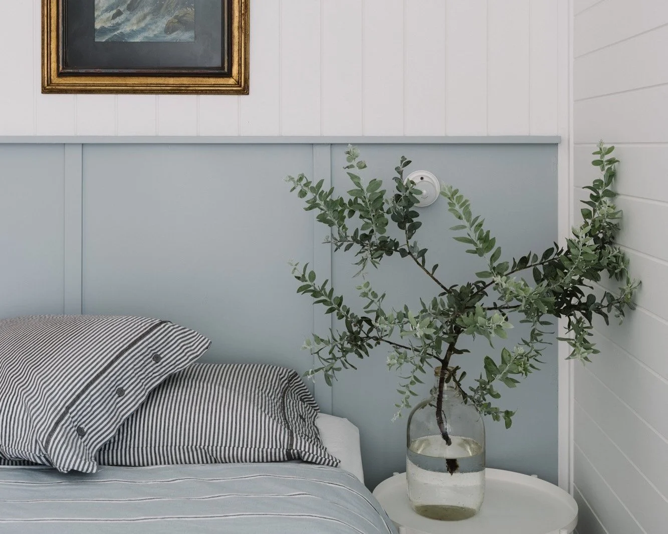 A bedroom corner featuring a bed with striped pillows, a light blue and white paneled wall, a framed painting above, a white side table, and a vase with a green leafy plant.