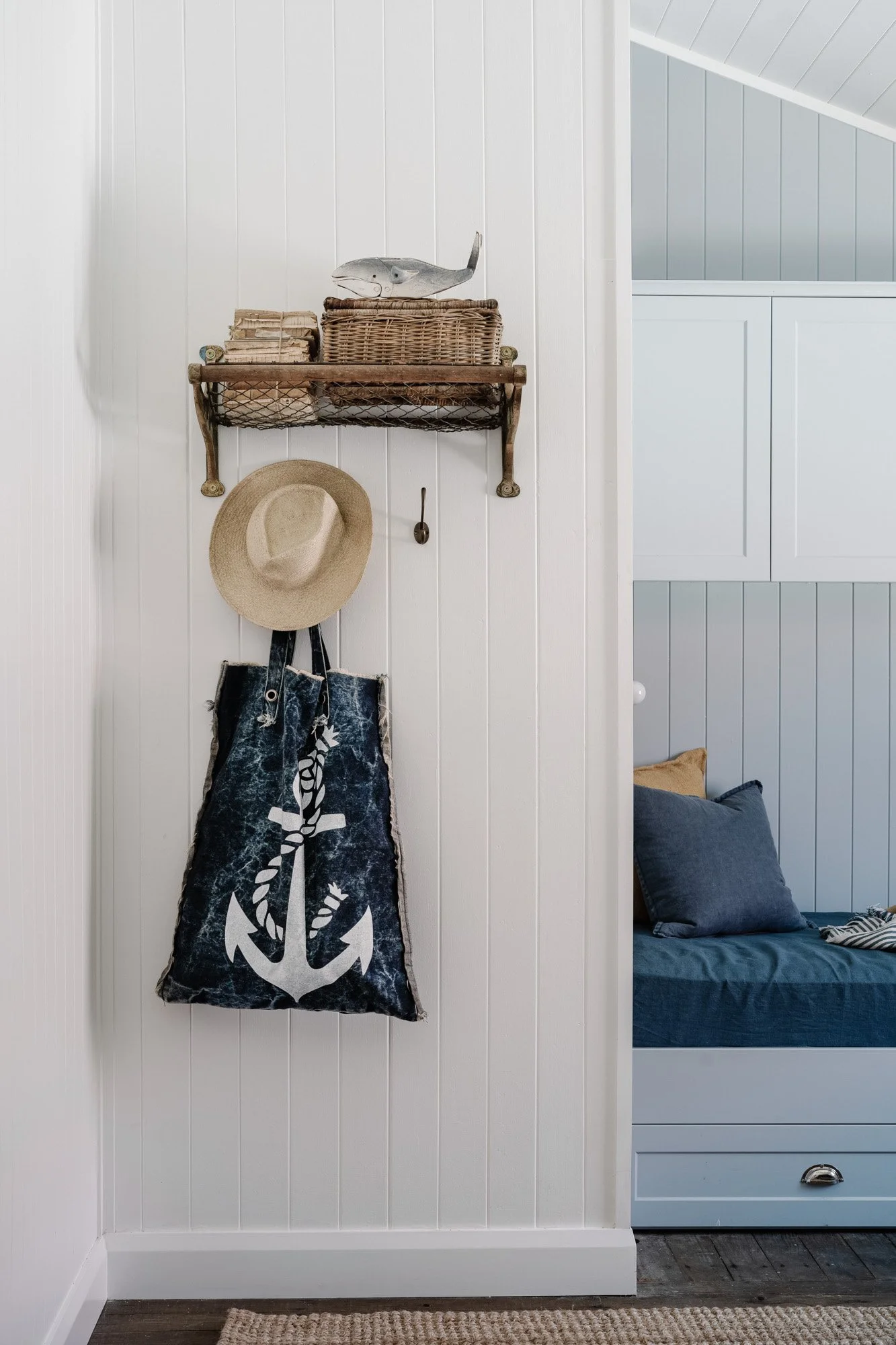 Coastal-themed wall decor with a wicker basket, a paper fish, a straw hat, and a bag with an anchor design, next to a bench with pillows in a bedroom.