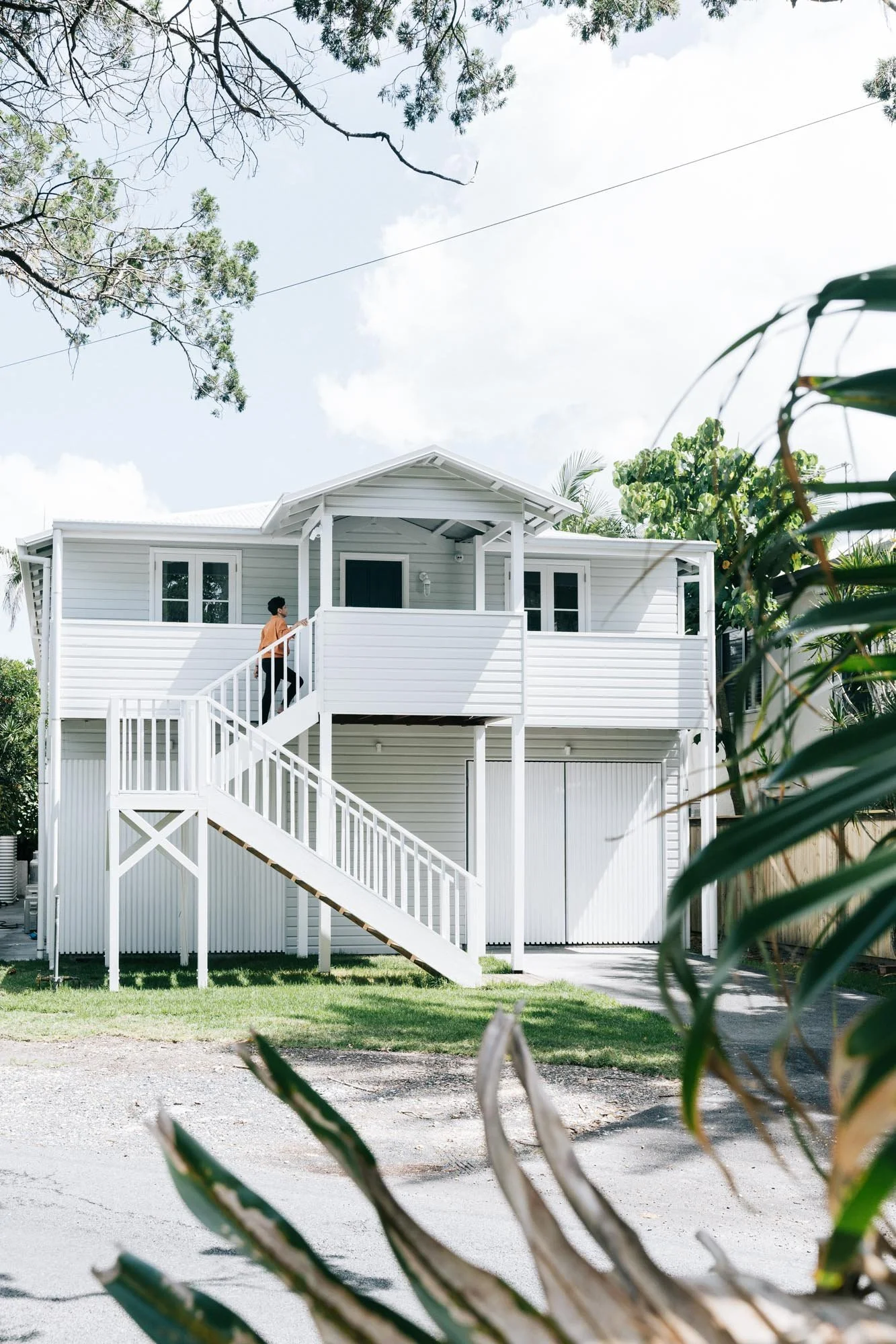 A white two-story house with an exterior staircase leading to the upper level, with a person standing on the balcony, surrounded by trees and greenery.