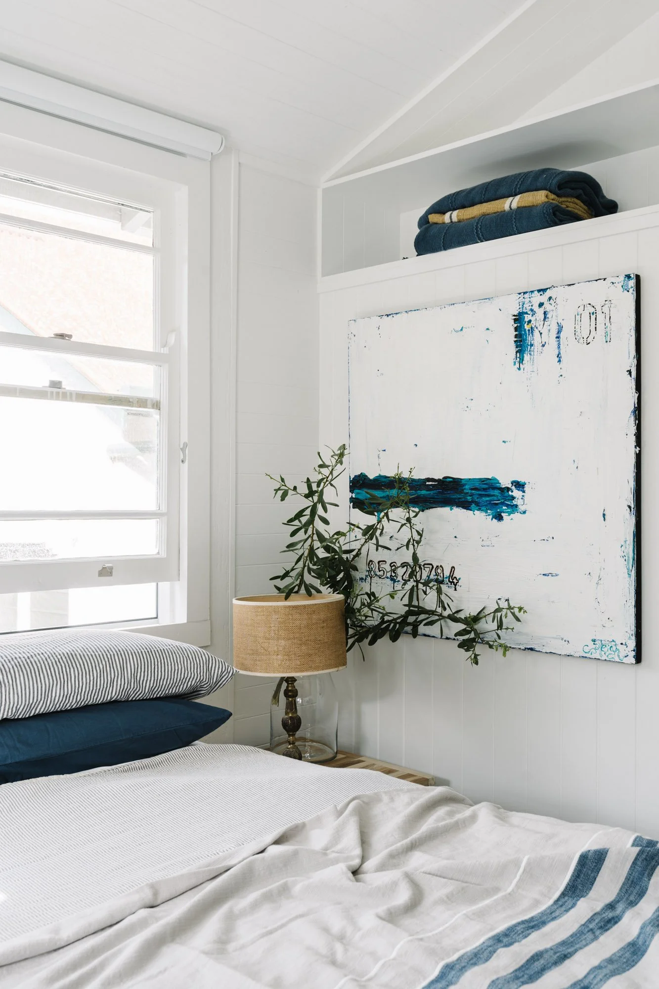 A cozy bedroom with a bed, striped pillows, a bedside lamp, a potted plant, and abstract artwork on the wall.