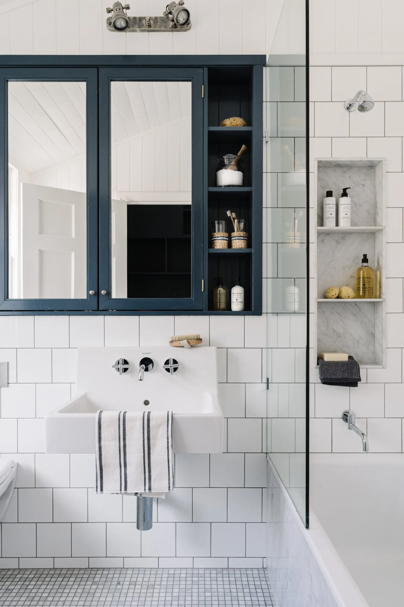 A bathroom with white tiled walls, a white sink with a striped hand towel, mirrored cabinet, floating shelves with toiletries, and a glass shower partition.