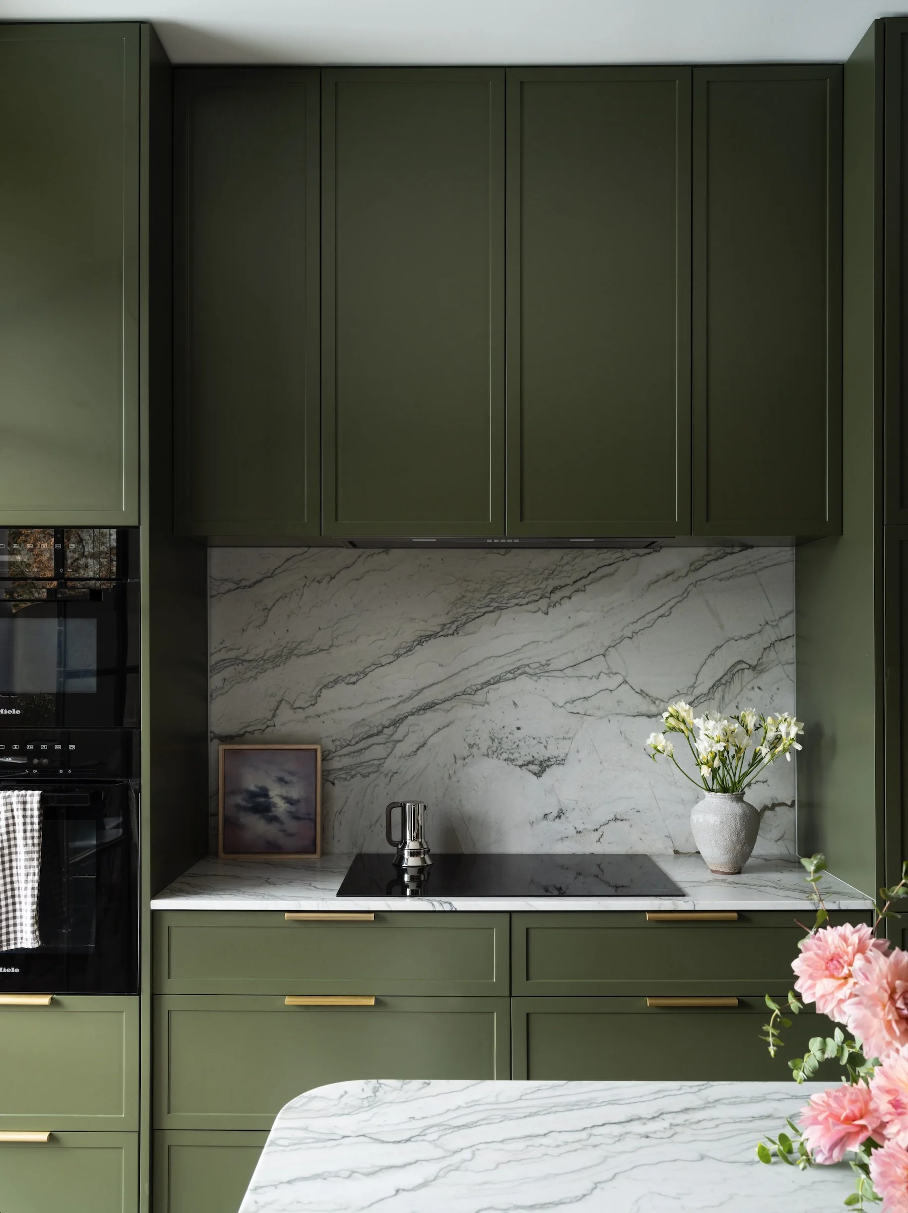 A modern kitchen with dark green cabinets, a white marble backsplash, and a countertop with a black induction cooktop. There is a small framed picture and a vase with white flowers on the counter, with a larger pink flower arrangement in the foreground.