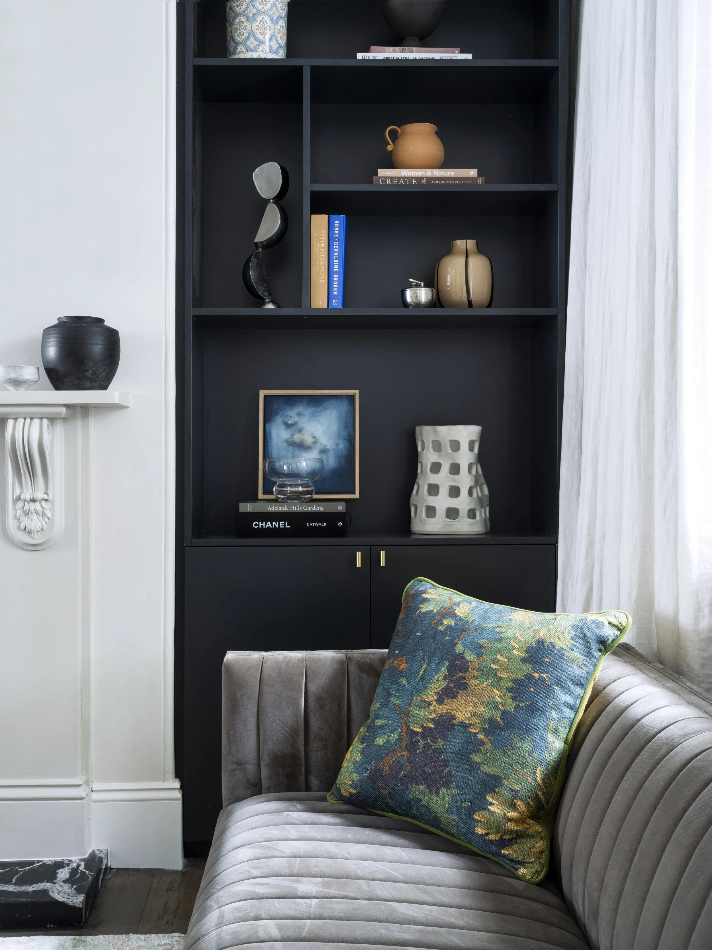 A living room corner with a beige velvet sofa with a green and blue floral pillow, a black bookshelf with decorative objects and books, a white ornate fireplace with black and white marble, and sheer white curtains.