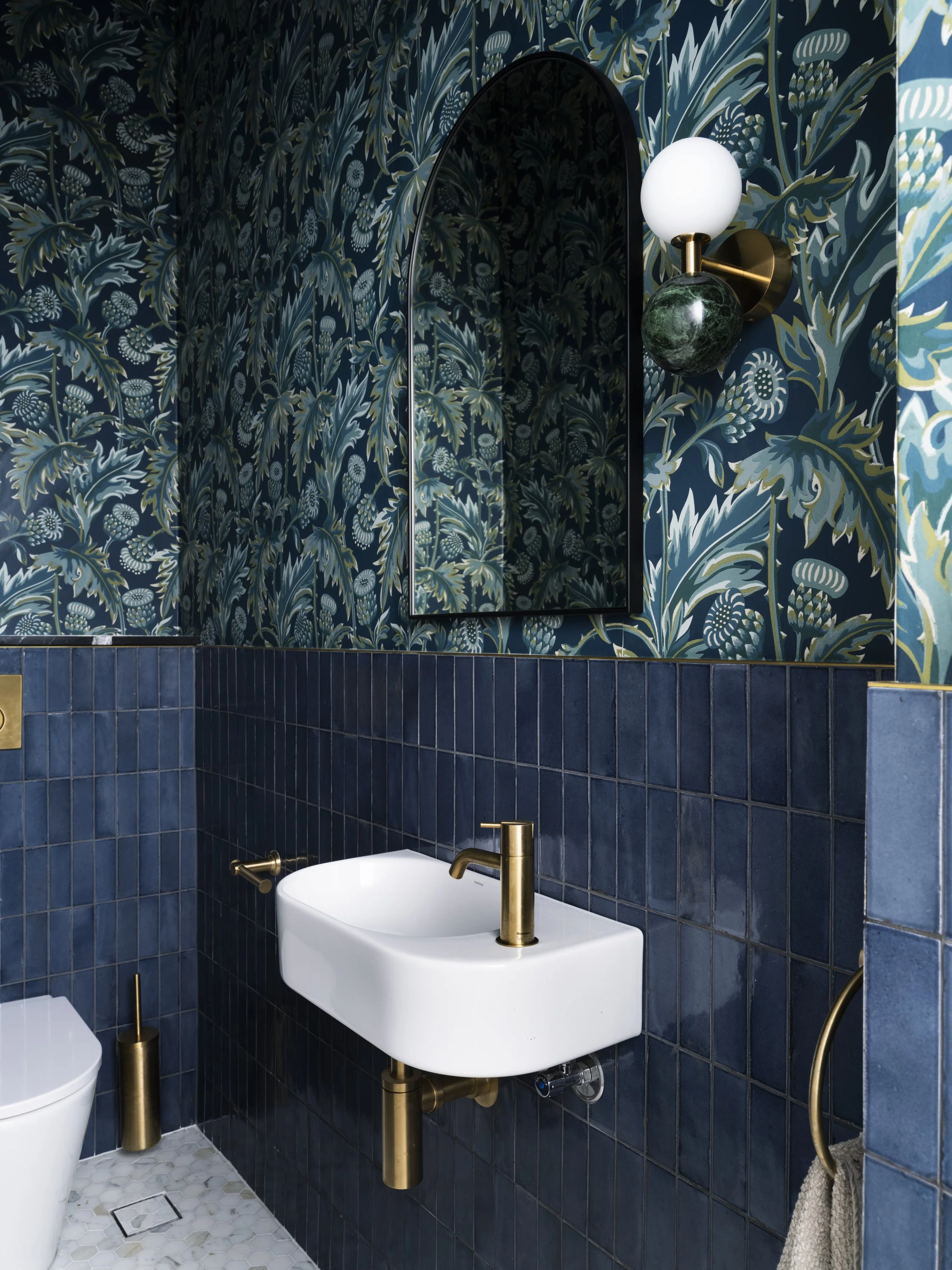 A bathroom with blue botanical wallpaper, dark blue tiled walls, a small white sink with a gold faucet, a oval mirror, a wall-mounted light fixture with a white globe and a green marble accent, a gold toilet paper holder, and a white toilet.