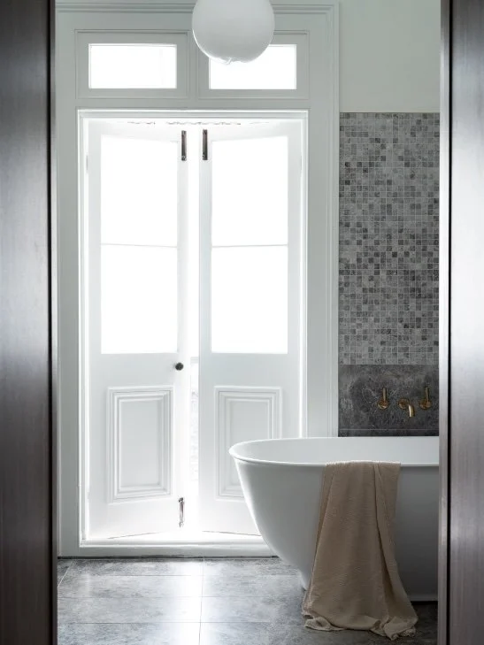 Bright bathroom with a white bathtub, a beige towel hanging over the edge, a tiled wall with small gray mosaic tiles, a door with glass panes, and gold fixtures.