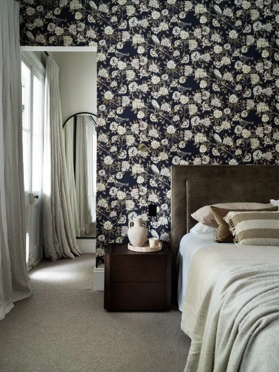 A bedroom corner featuring a floral wallpapered accent wall, a large bed with beige bedding and pillows, a dark wooden nightstand with a white vase and candles, and a window with sheer white and heavier white curtains.