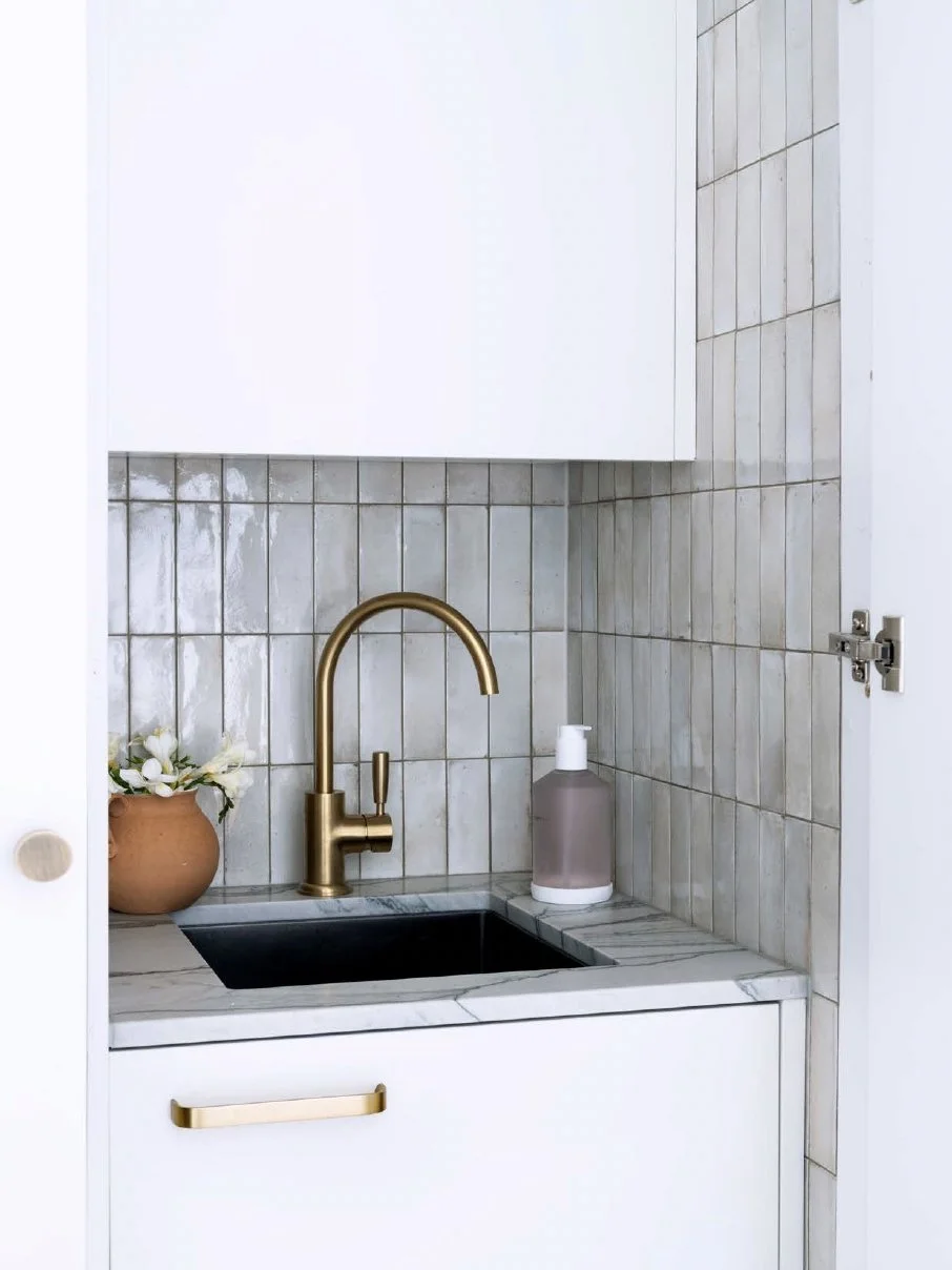 Small kitchen sink with gold faucet in a minimalist style, beige backsplash tiles, white cabinets, a small potted plant with white flowers, and a soap dispenser.