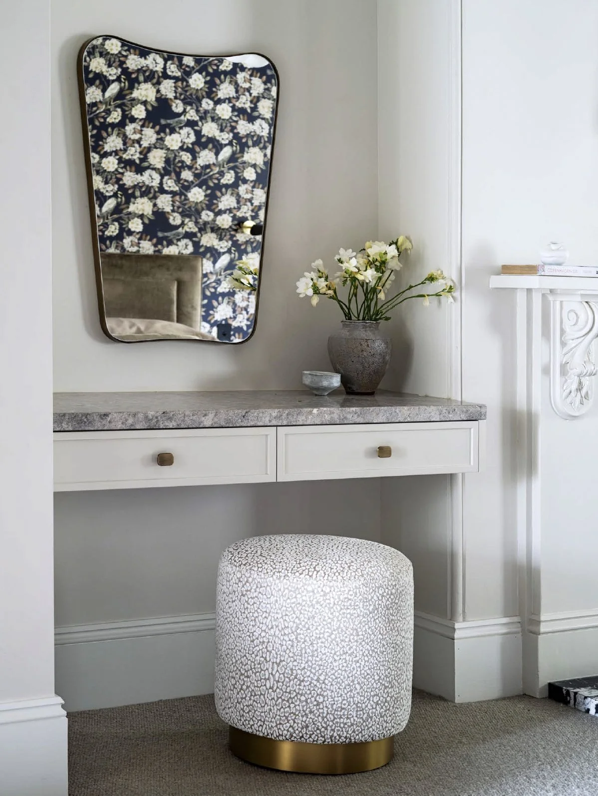 A white vanity table with two drawers and a marble top, accompanied by a round, patterned ottoman with a gold base. A gray vase with white flowers and a small bowl sit on the table, and a large, irregularly shaped mirror with a gold trim hangs above. Part of a floral patterned wallpaper is reflected in the mirror.