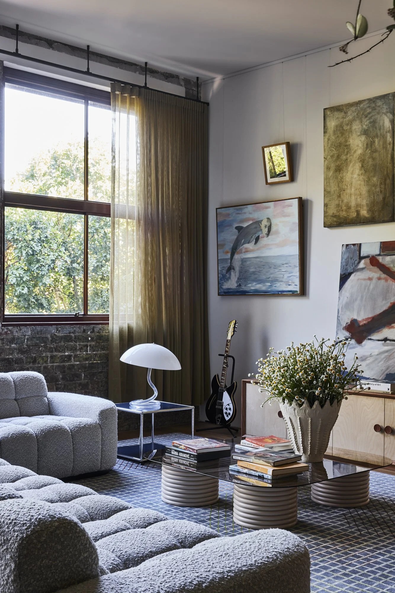Living room with large window, beige sofa, glass coffee table with books, white lamp, guitar, and artwork on white walls.