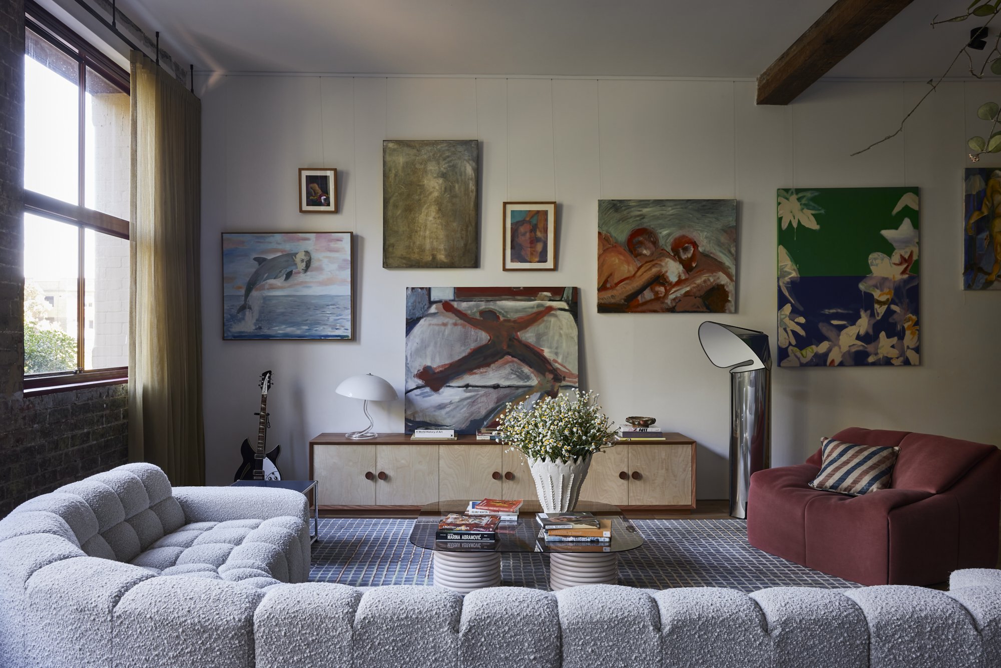 Living room with a white textured sofa, a red velvet armchair, a glass coffee table with books and a vase of flowers, and various paintings on the wall. There's a window with beige curtains, a guitar, and contemporary lamps.