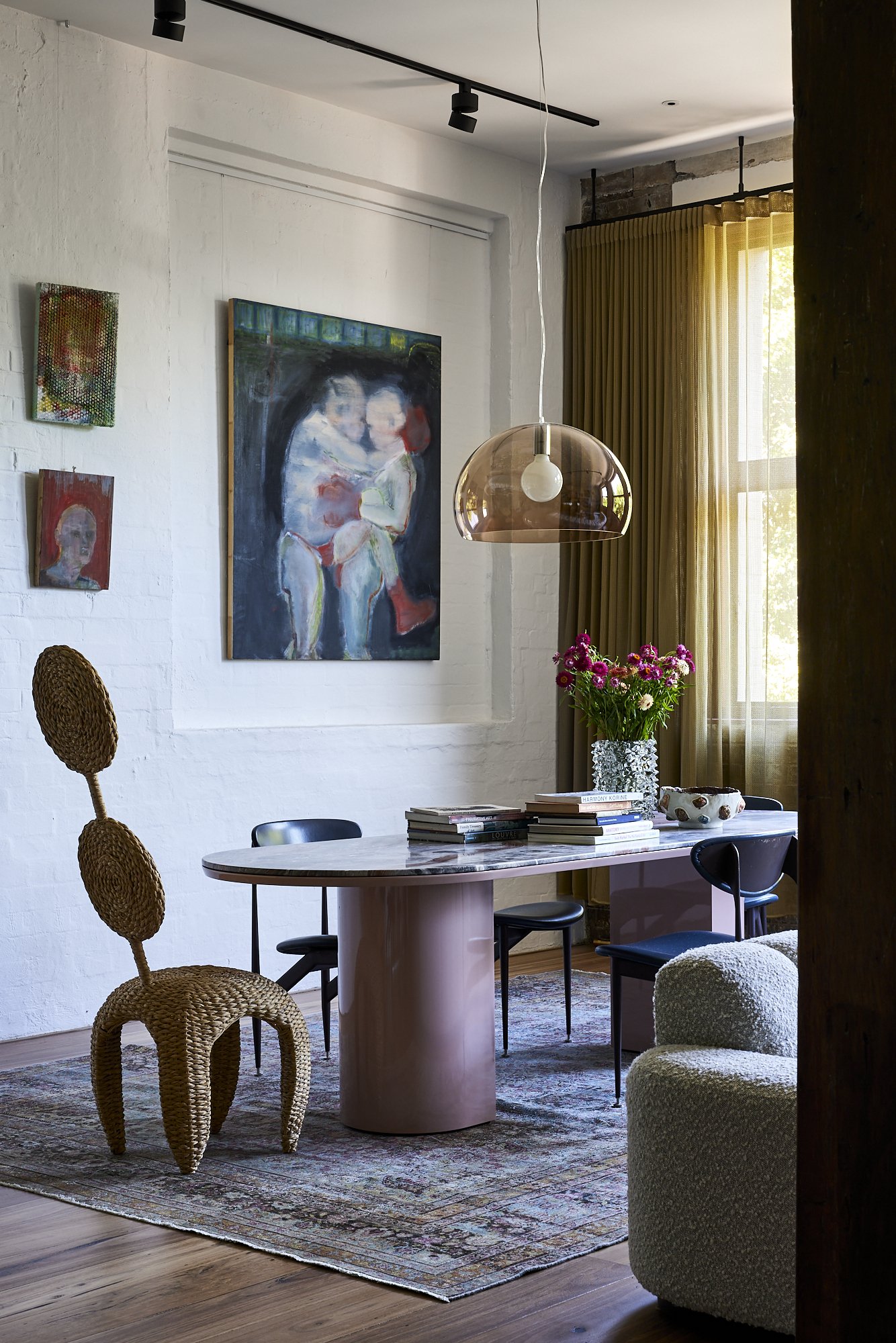 A dining room with a curved marble table, surrounded by black chairs, a woven chair, and a plush cream armchair. There is a vase of pink flowers on the table, a stack of books, and artwork on the white brick wall. A large window with yellow curtains lets in natural light.