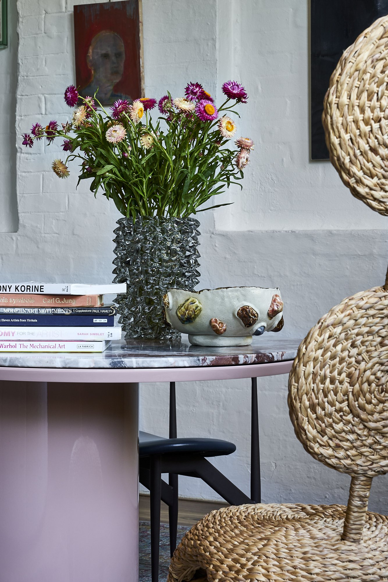 A pink marble-top table with a glass vase filled with pink and white daisies, stacked books, a ceramic bowl with colorful bumps, a rattan chair, and framed art on a white brick wall.